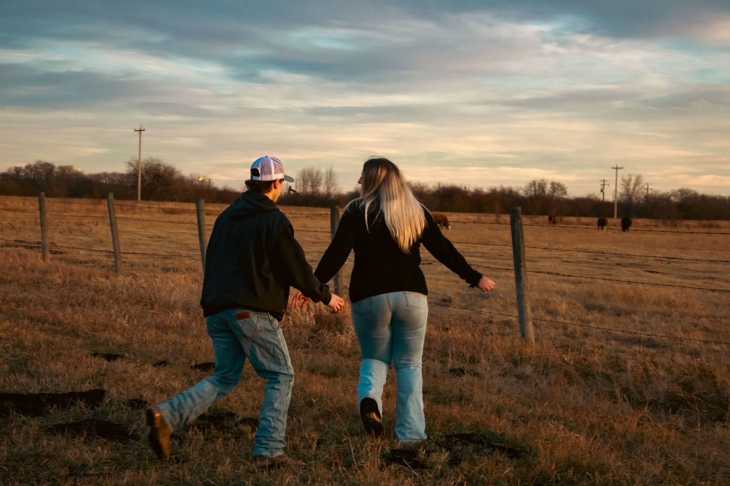 Couple holding hands walking along a fence in a rural field at sunset.