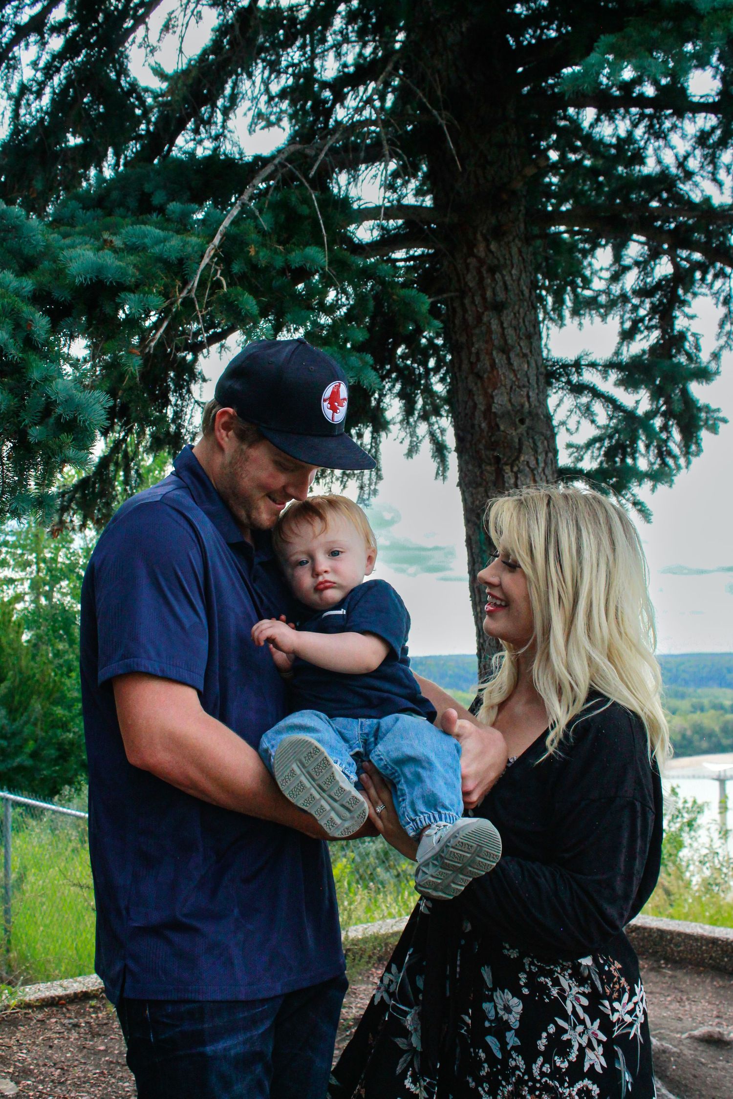 Happy young family with blonde mother, father in cap, and baby boy under pine tree.