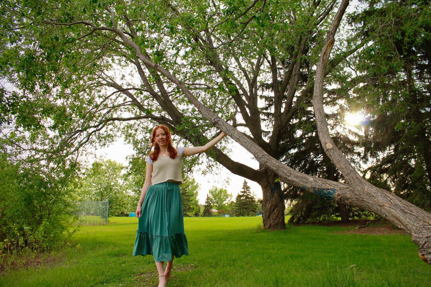 Red-haired woman in teal midi skirt walks barefoot on grass while touching a leaning tree at sunset.