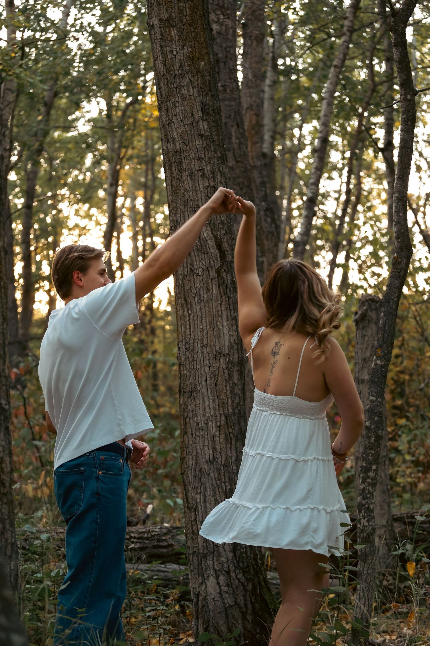 Couple dancing in a sunlit autumn forest, man in white shirt spinning woman in flowing white dress.