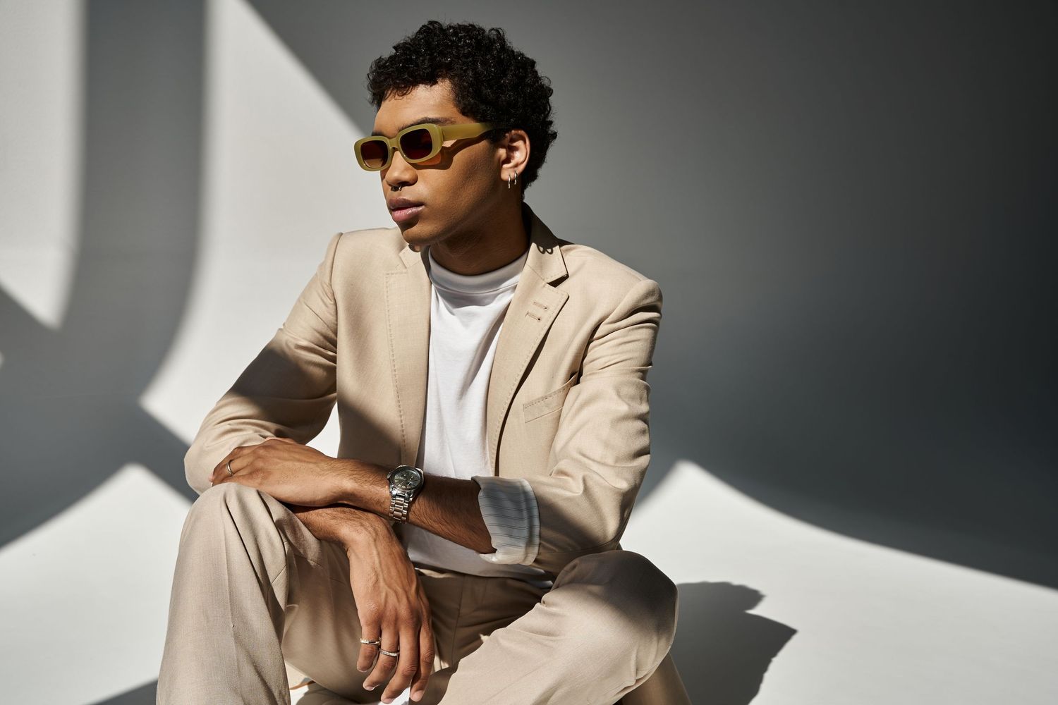 Stylish person in beige blazer and sunglasses poses against gray wall in modern fashion photoshoot.