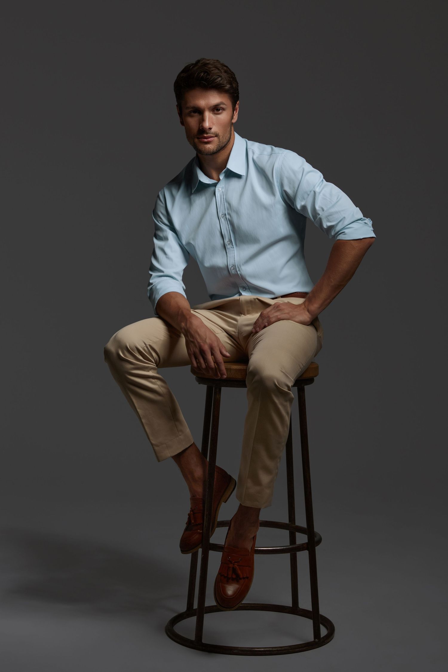 Person in light blue button-down shirt and khaki pants sits casually on dark wooden rocking stool against gray backdrop.