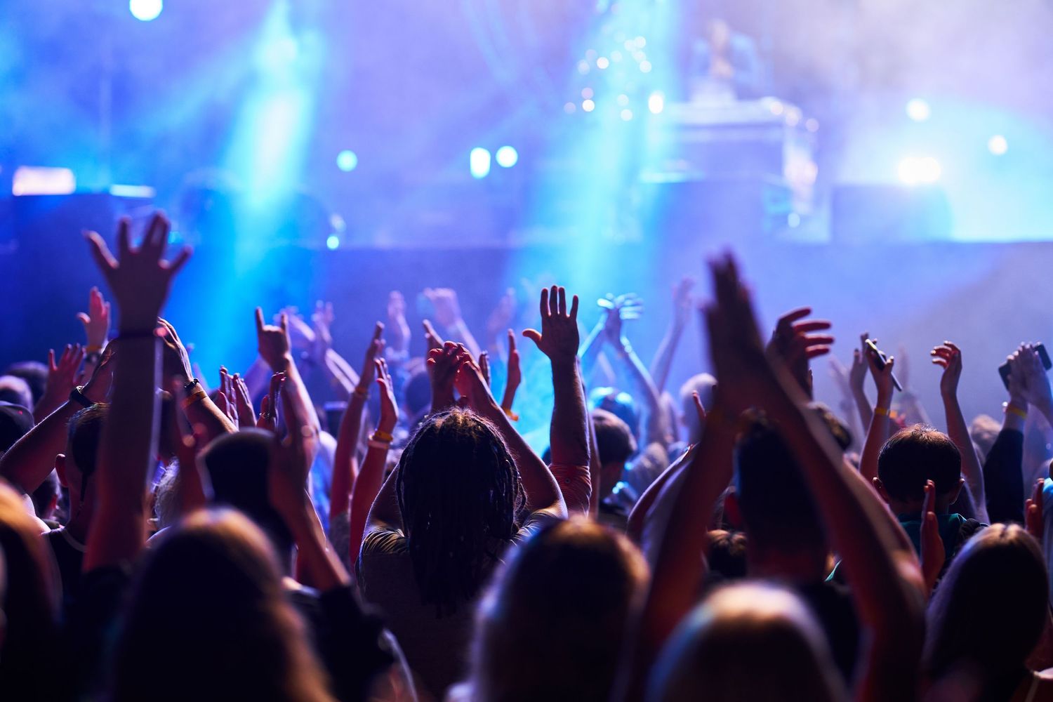 Silhouetted crowd with raised hands at a concert illuminated by bright blue stage lighting and spotlights.