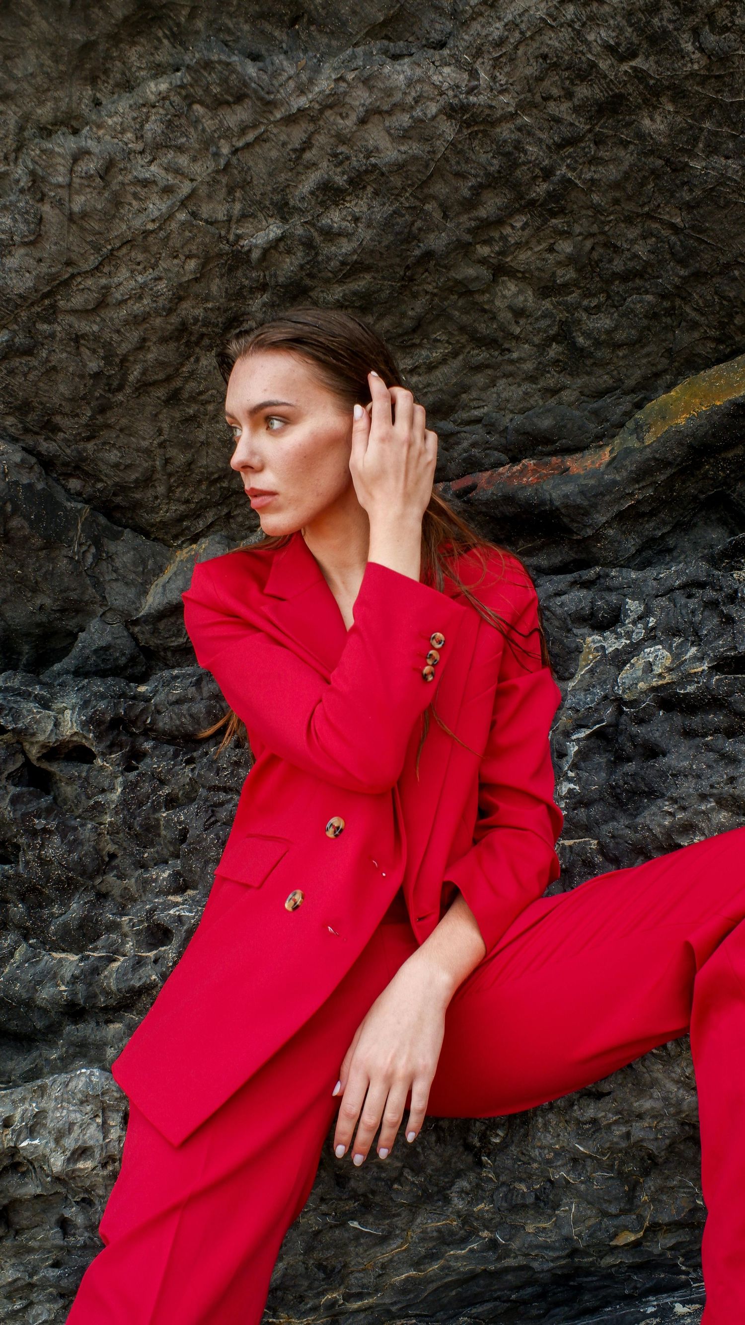 Fashion model in vibrant red suit poses elegantly against textured rock wall in artistic photo series.