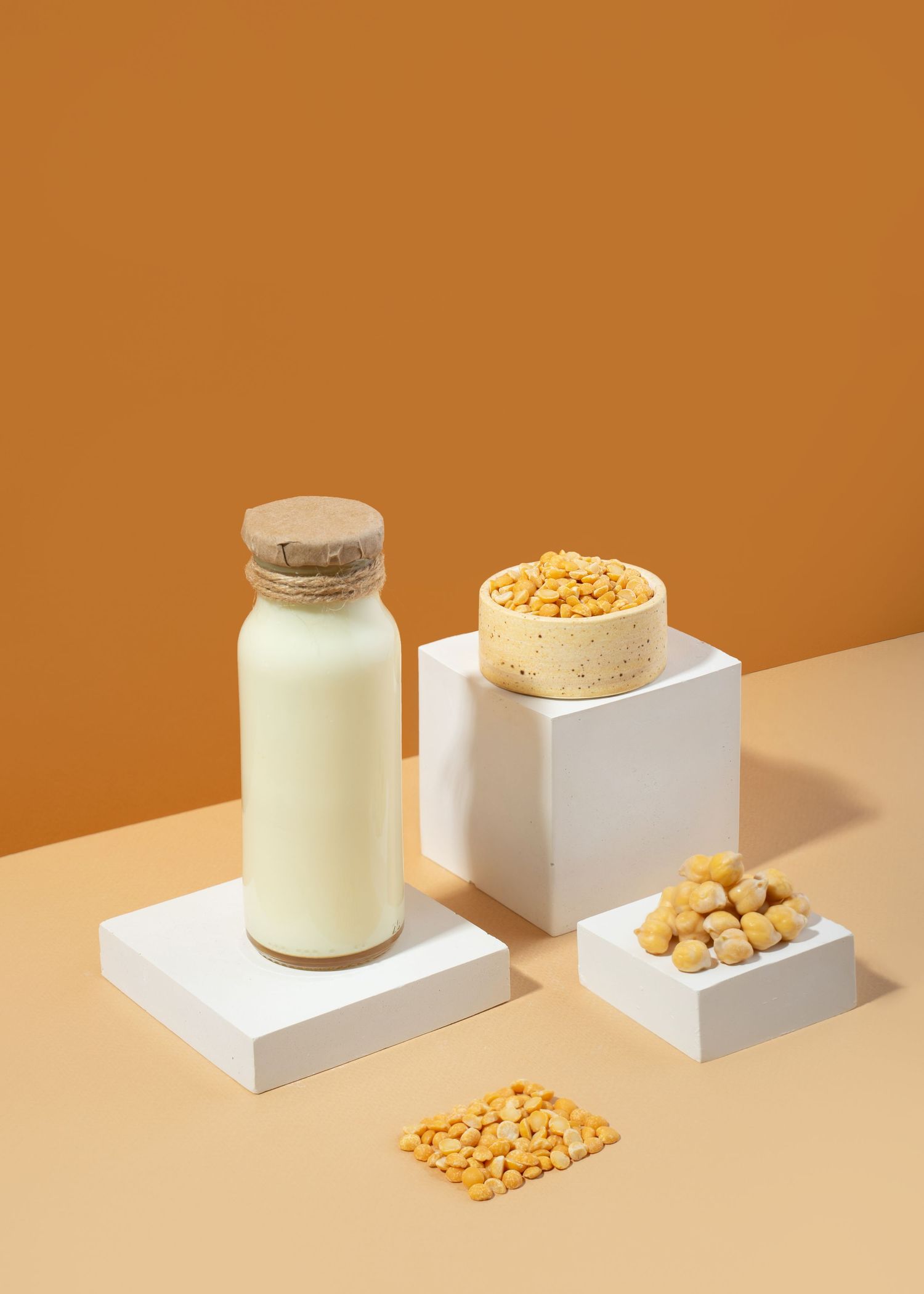 A minimalist composition showing glass jars of soy milk and soybeans on white platforms against an orange background.