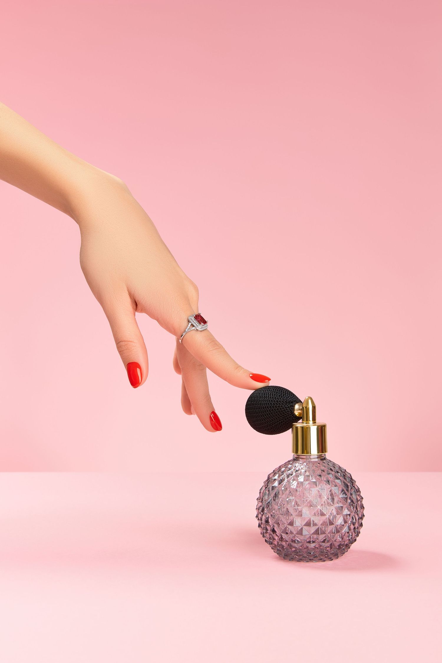 Hand with red nail polish placing a decorative purple glass vase against a pink background in a minimalist setup.