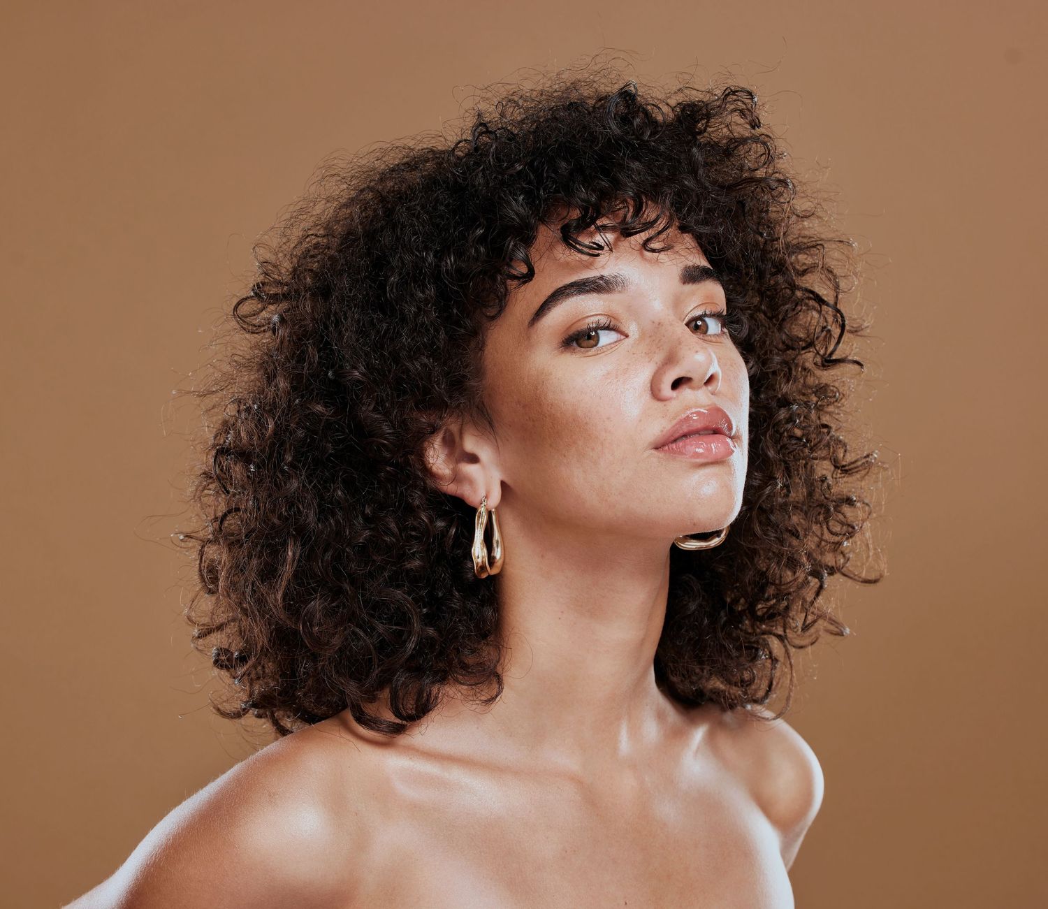 A model with curly dark hair and hoop earrings poses against a neutral brown backdrop.