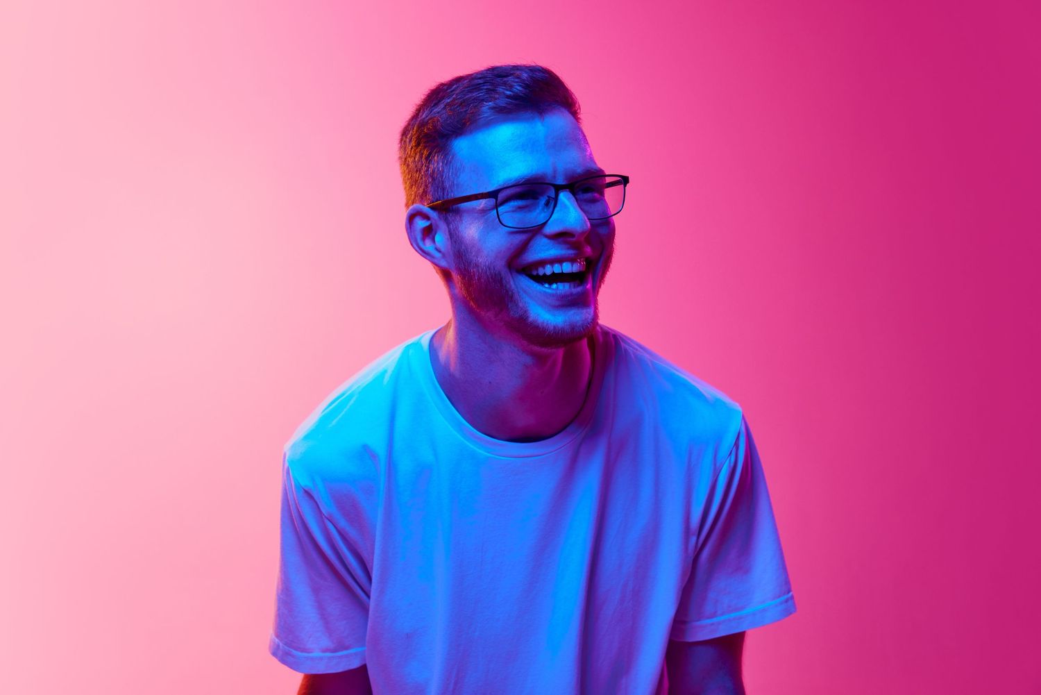Person in white t-shirt laughing against vibrant pink and blue lighting background.