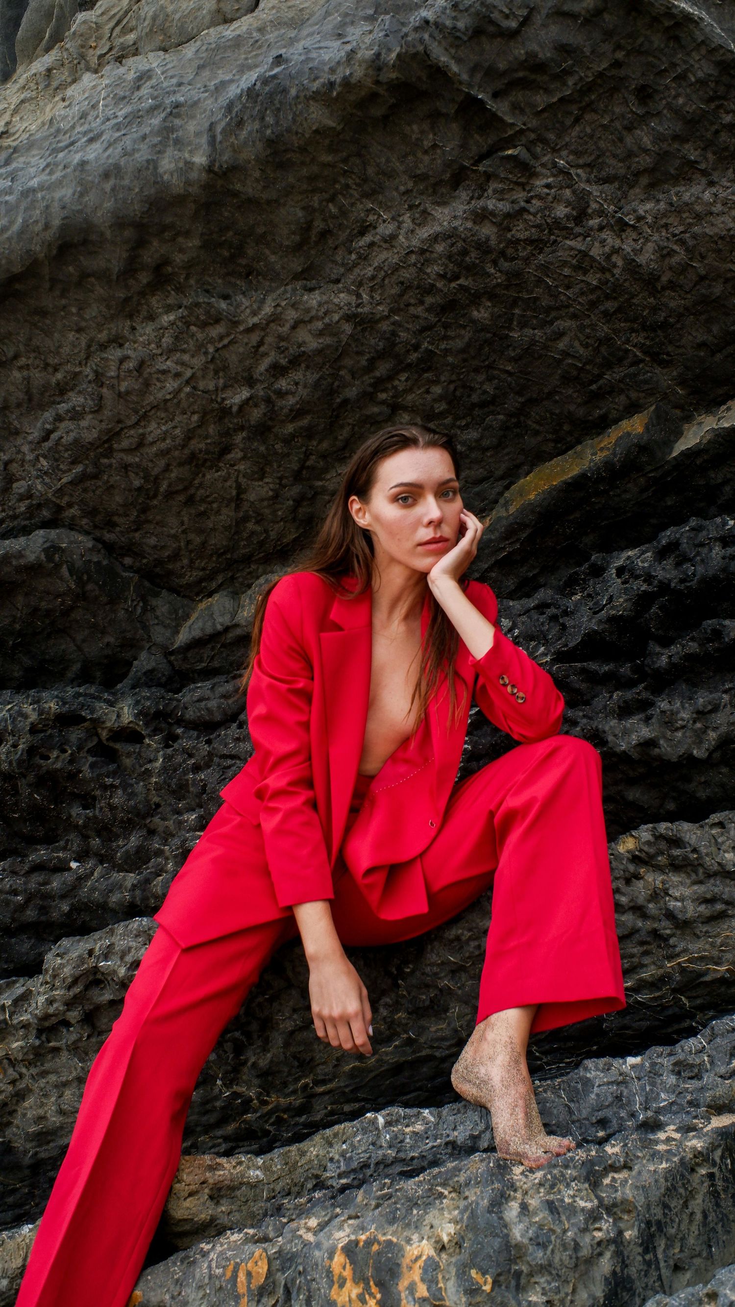 A figure in a striking red blazer poses dramatically against dark rocky cliffs in a fashion-forward photoshoot.