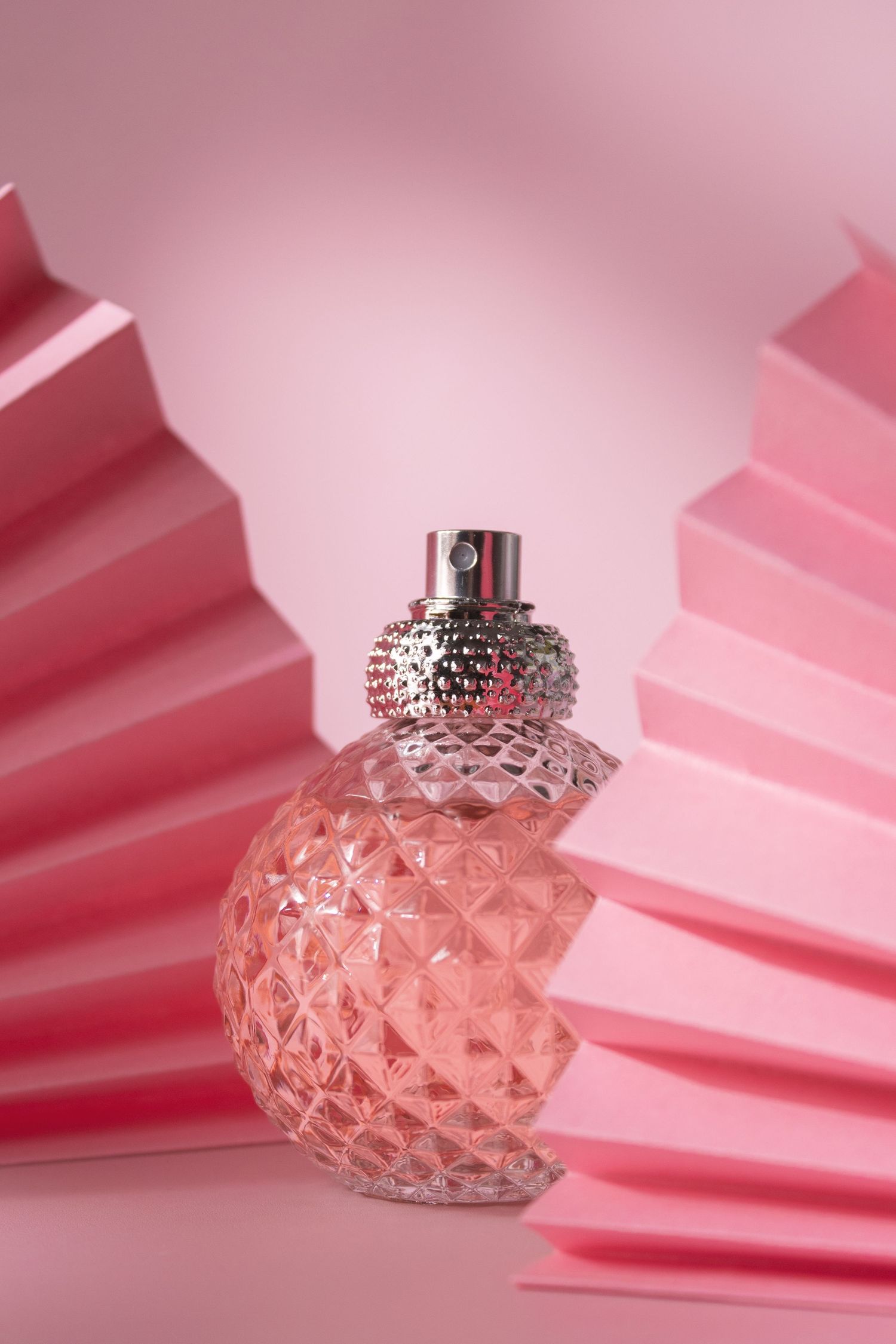 Pink glass perfume bottle with diamond pattern texture displayed against folded pink paper background.