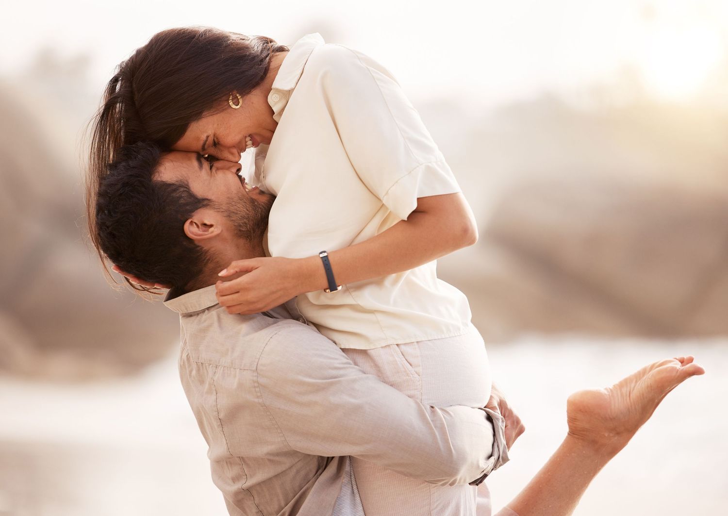A romantic couple in white embraces on a beach at sunset with soft golden light illuminating their silhouettes.