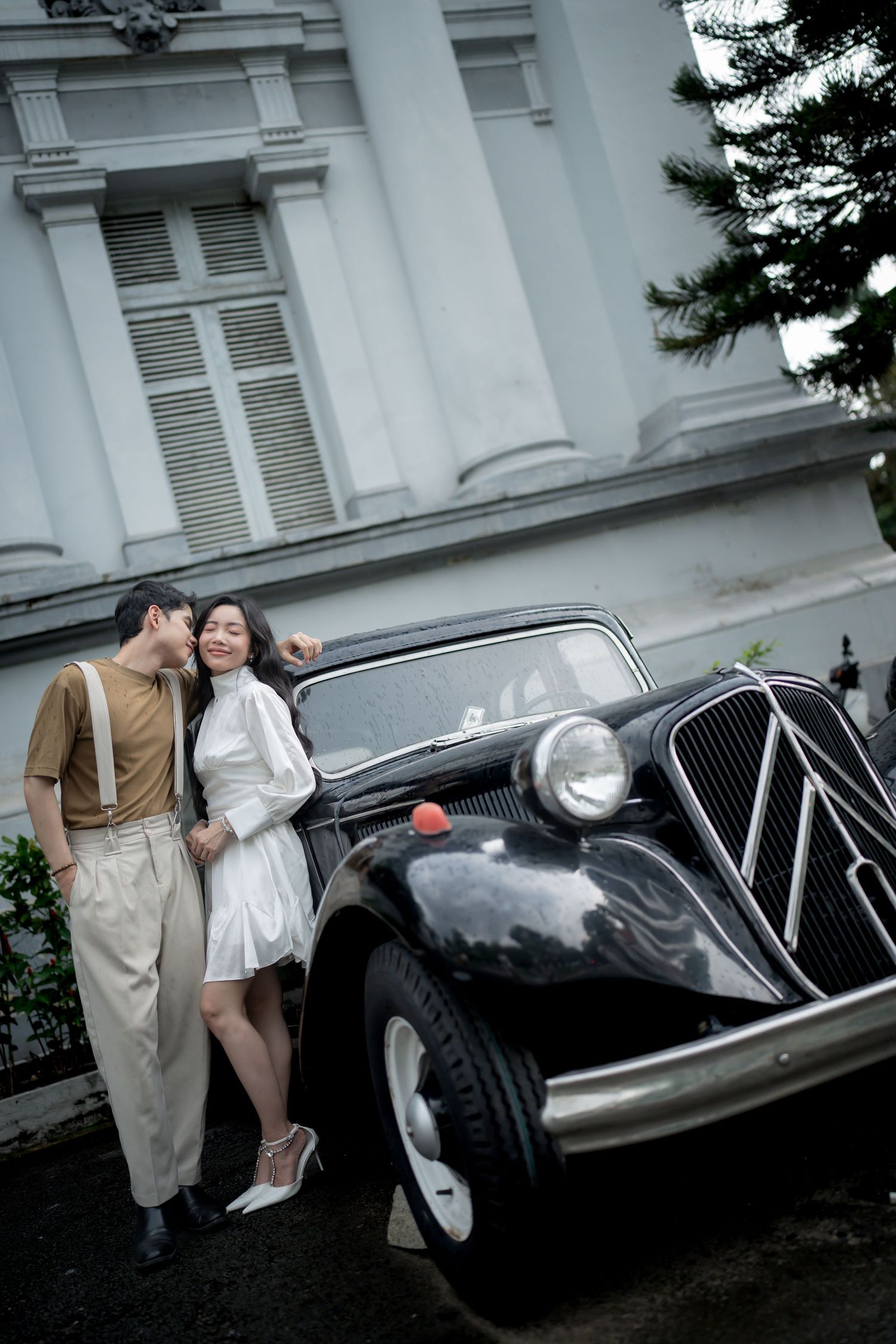 A classic black vintage car is showcased with a couple in retro outfits against a white colonial building.