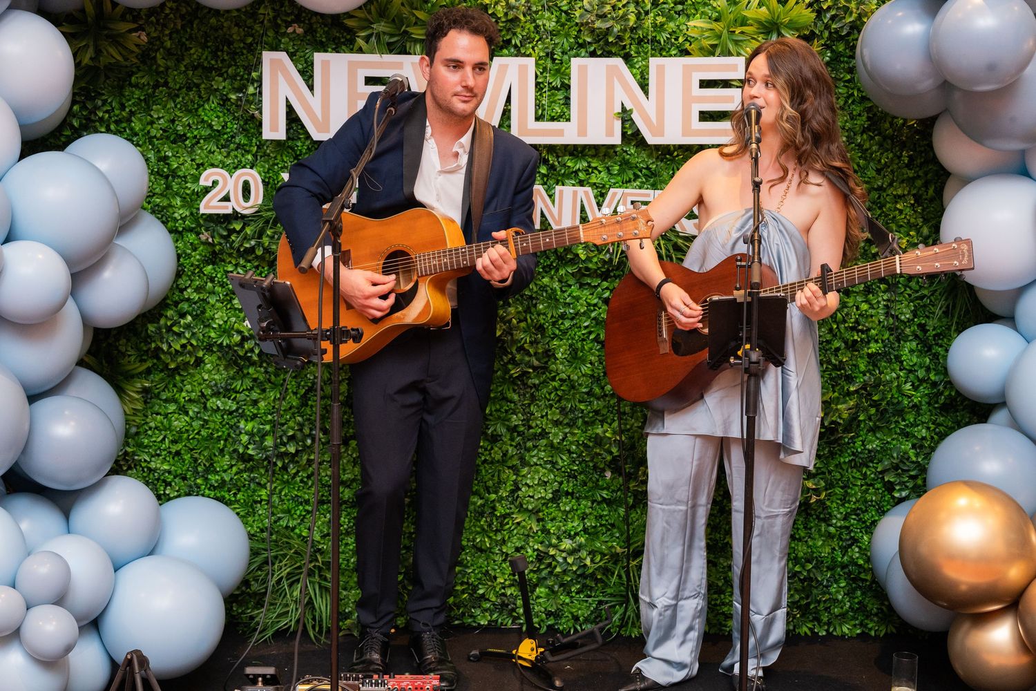 Two musicians playing acoustic guitars on stage at a New Line anniversary event with balloon decorations.