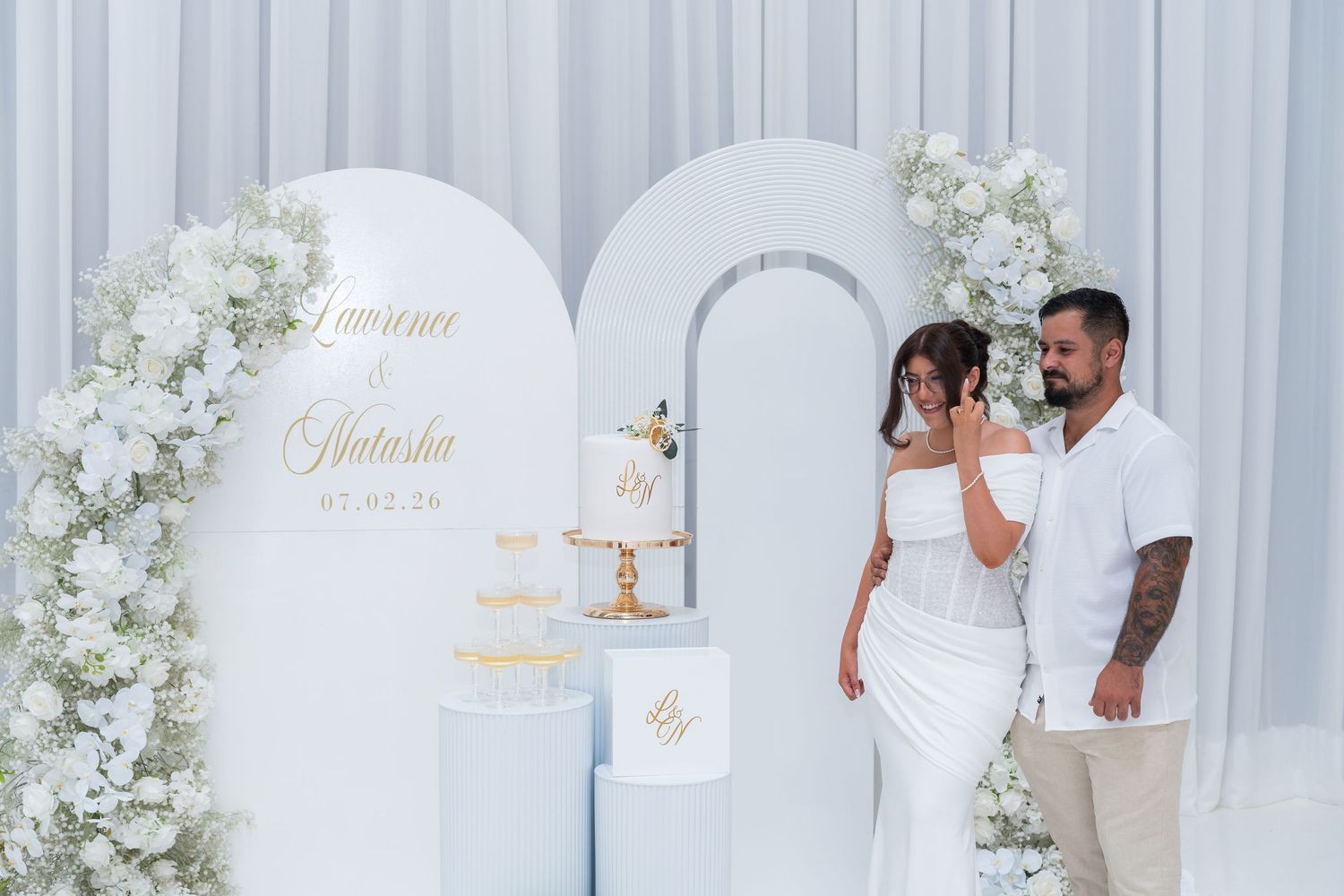 Elegant white wedding backdrop with floral arch, tiered cake, and couple in formal attire celebrating their special day.