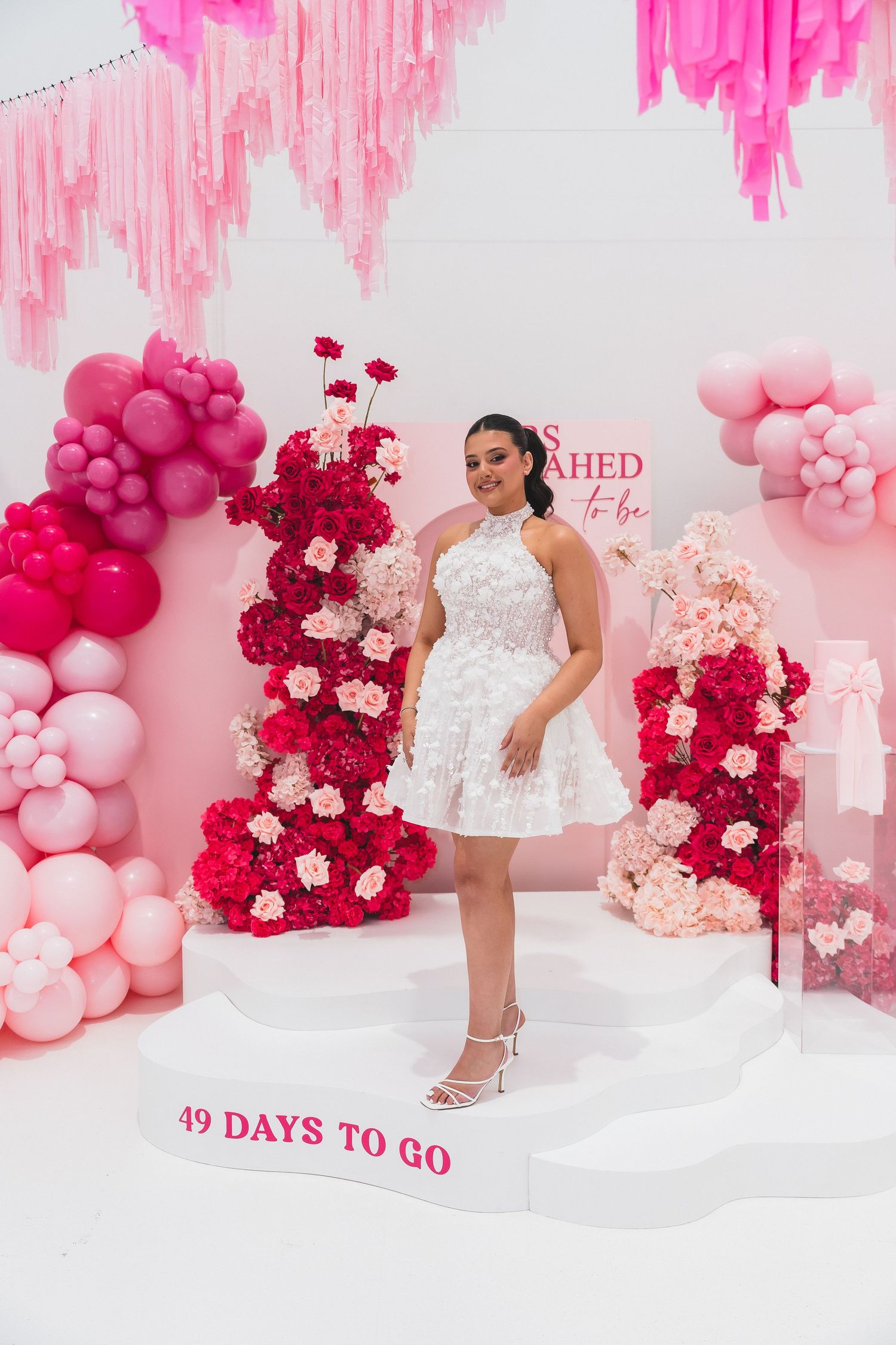 Bride-to-be in white dress posing at pink floral bridal shower with 49 Days To Go sign.