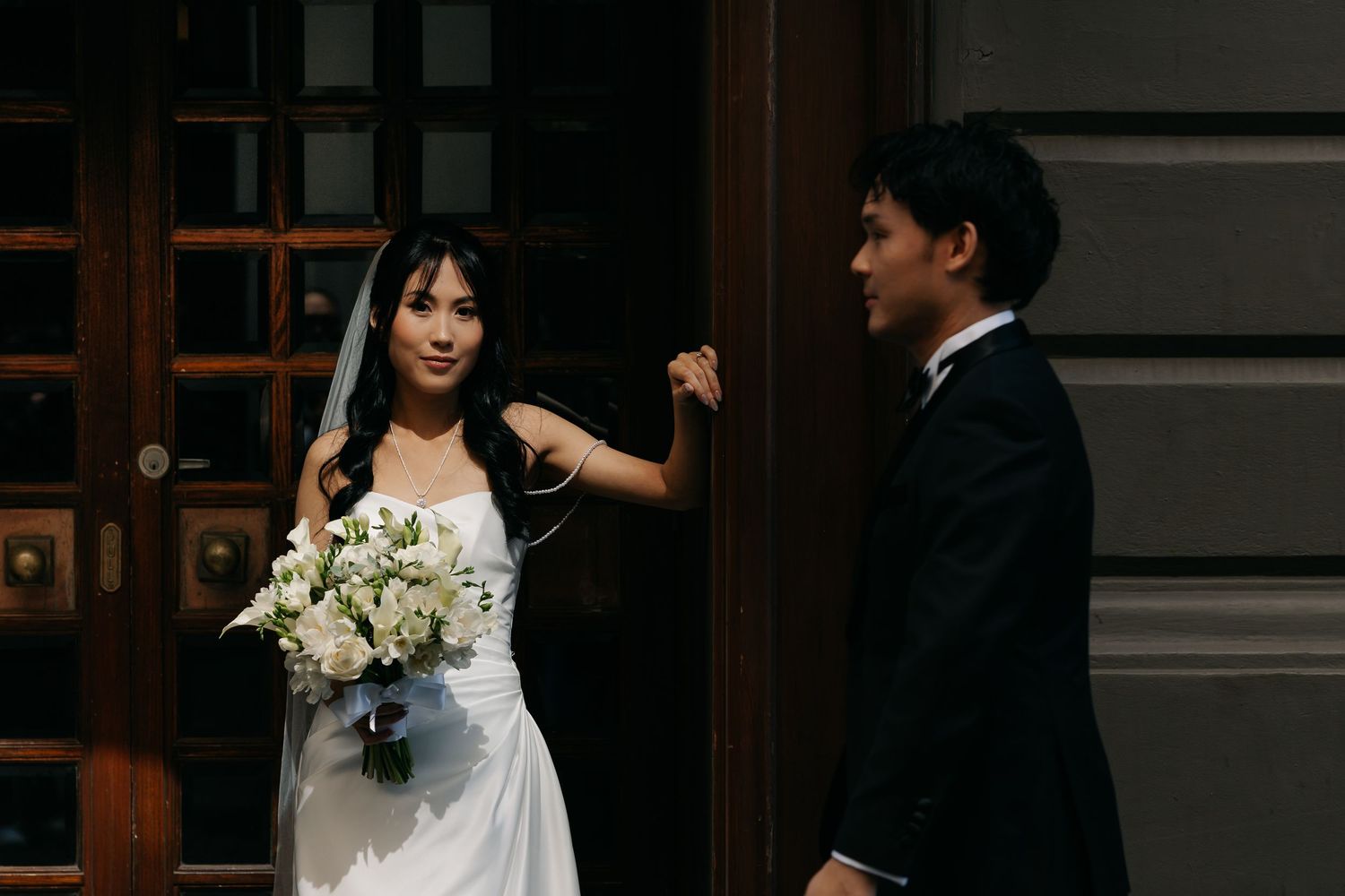 Bride in white gown holding bouquet stands with groom in black suit by ornate wooden doors.