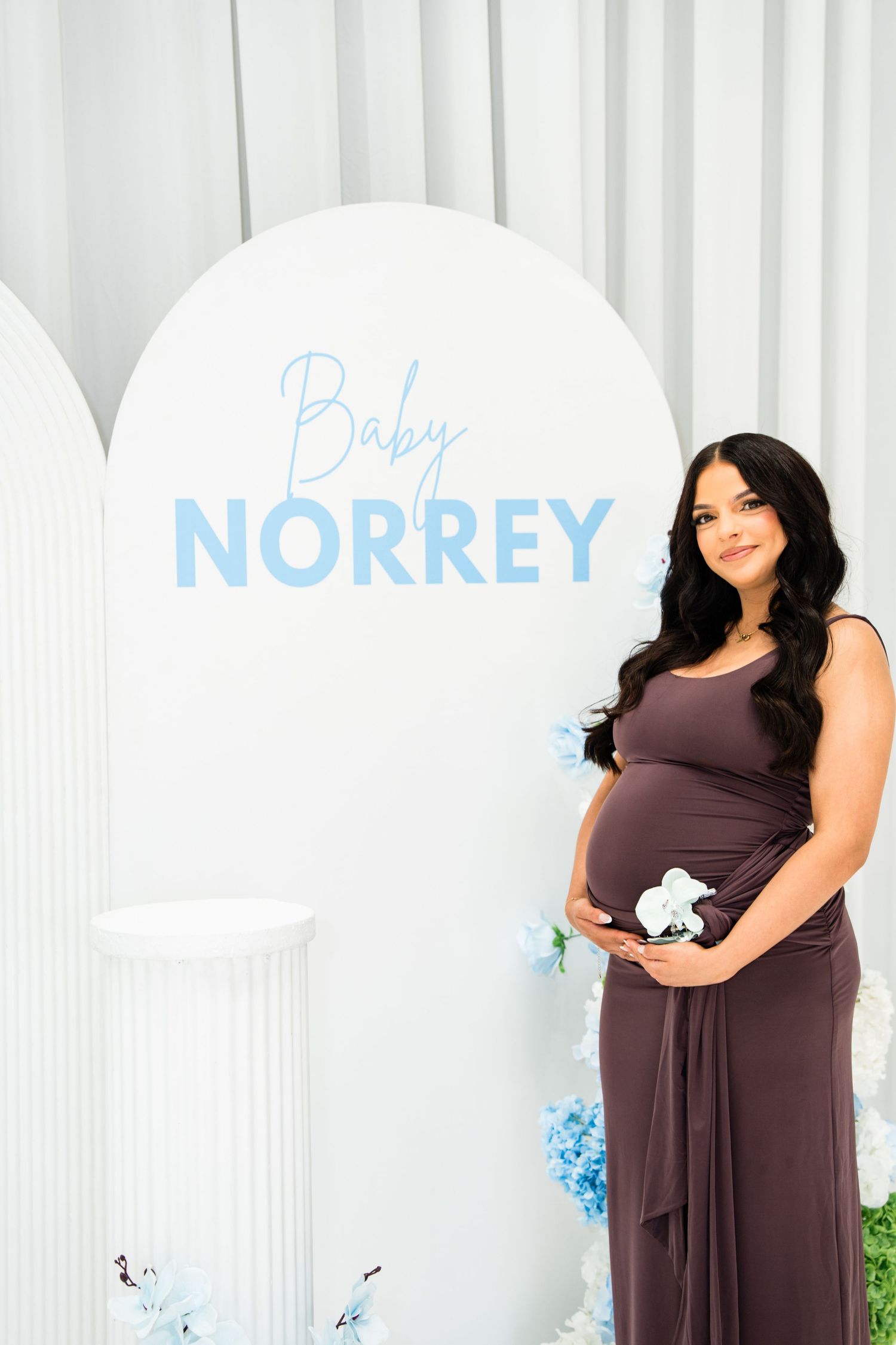 Pregnant woman in brown dress holding belly at Baby Norrey gender reveal shower with blue floral decor.