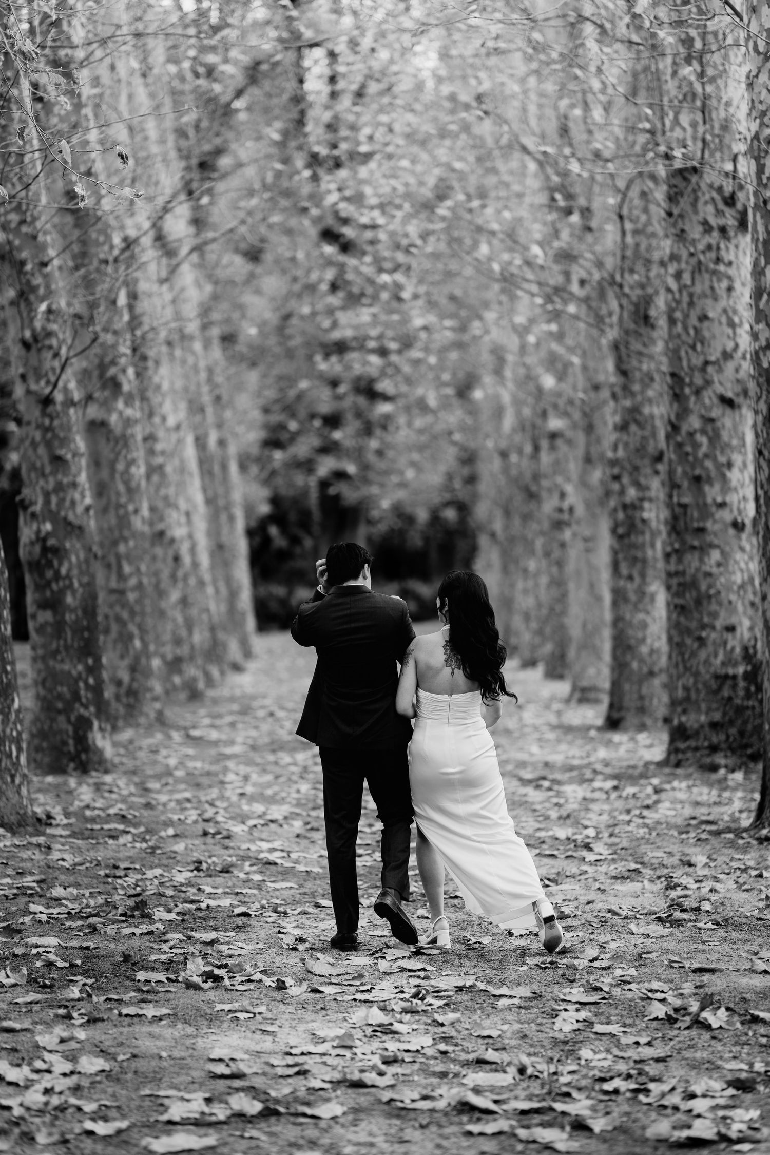 Black and white photo of bride and groom walking hand in hand through a tree-lined autumn path.