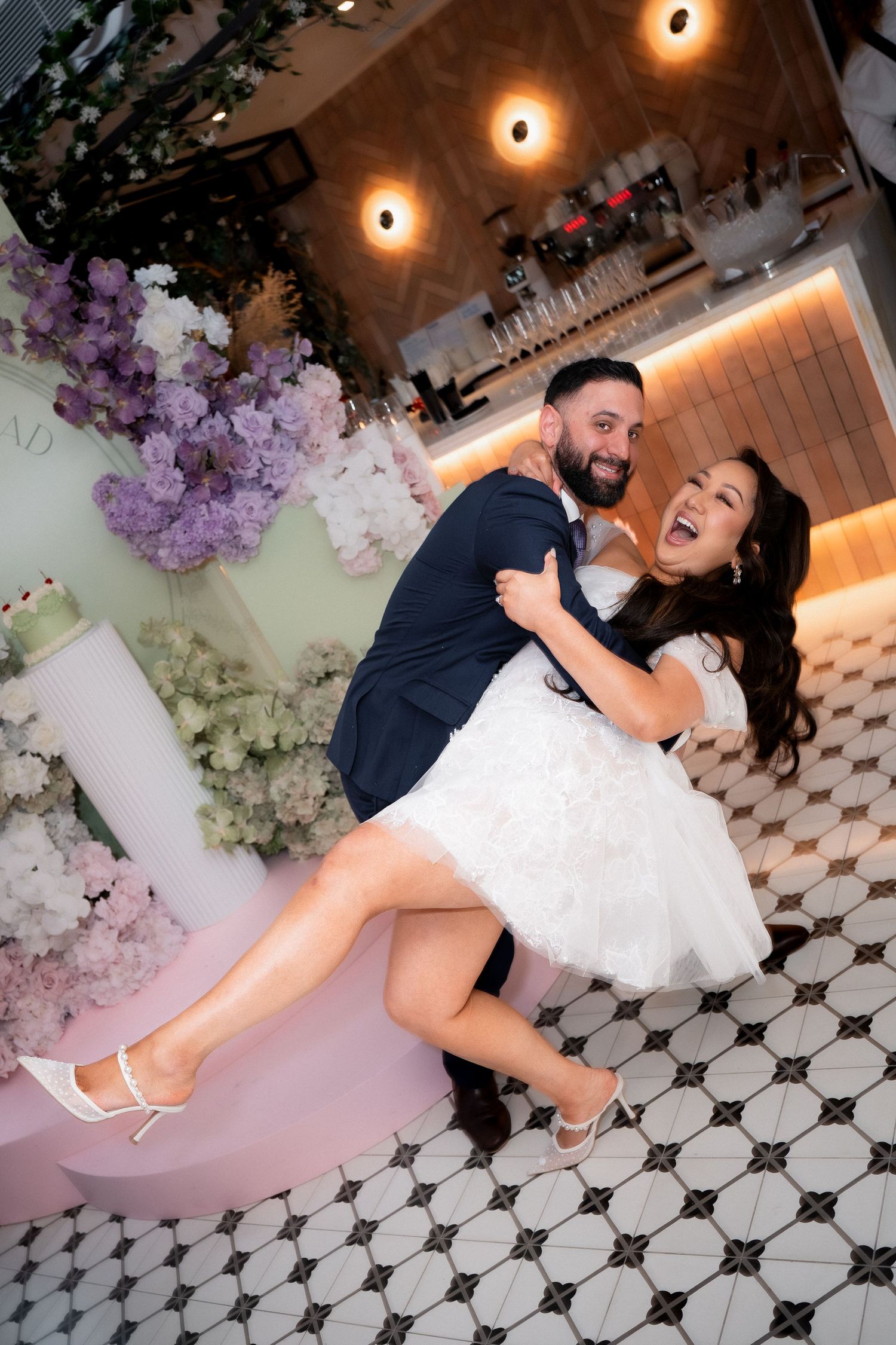 Joyful bride in white mini dress lifted by groom in navy suit, surrounded by pink and white floral arrangements.