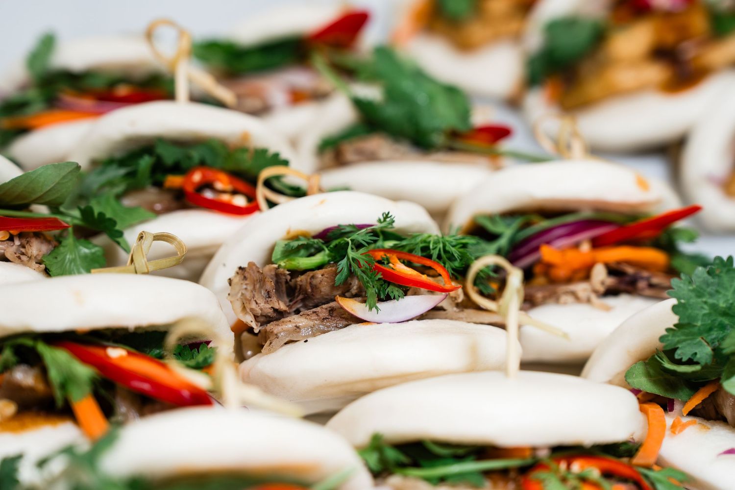 Steamed bao buns filled with pulled pork, fresh herbs, red chili, and colorful vegetables, served as appetizers.