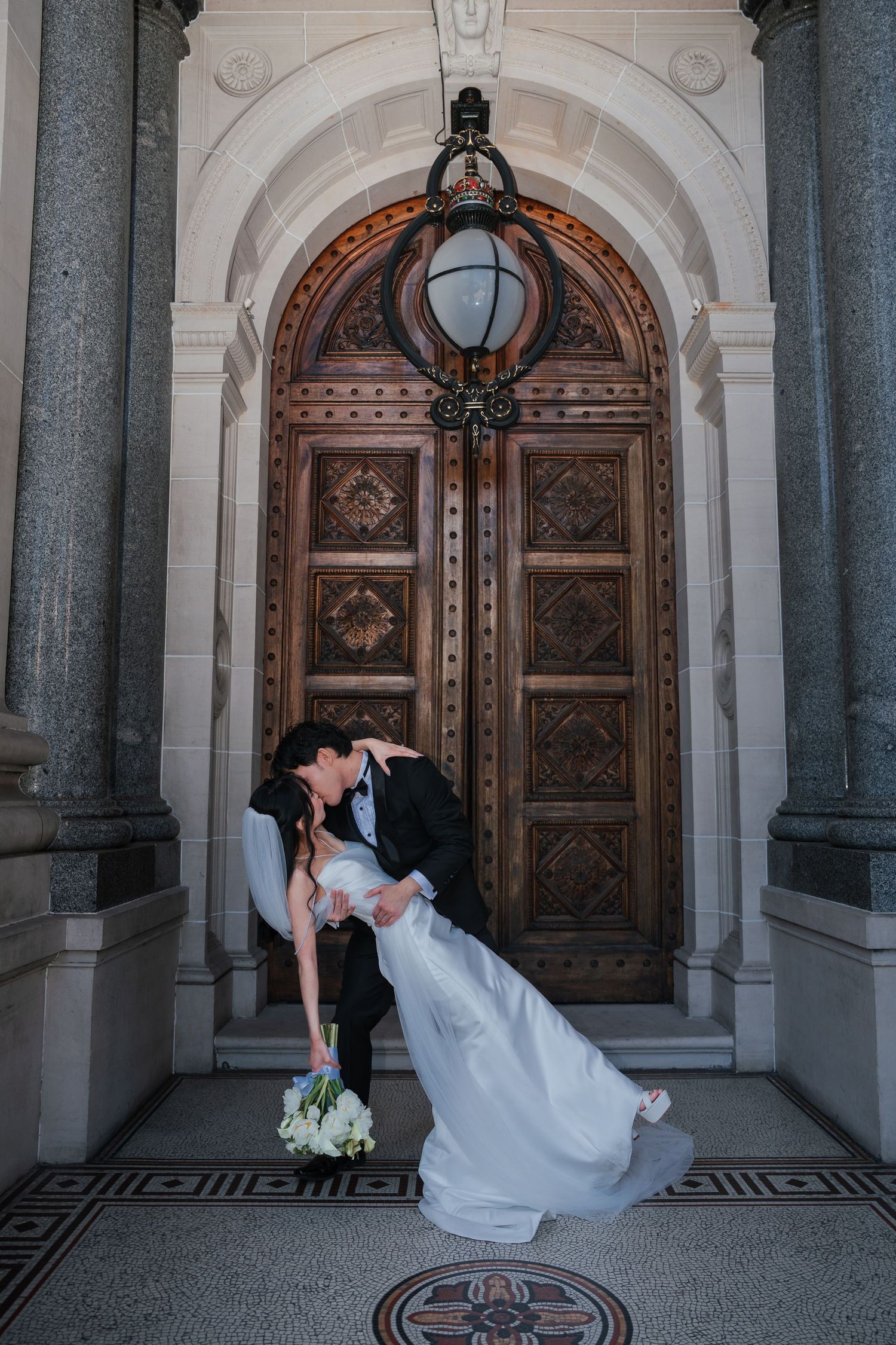 Groom dips bride in kiss before ornate wooden doors of grand building on wedding day.