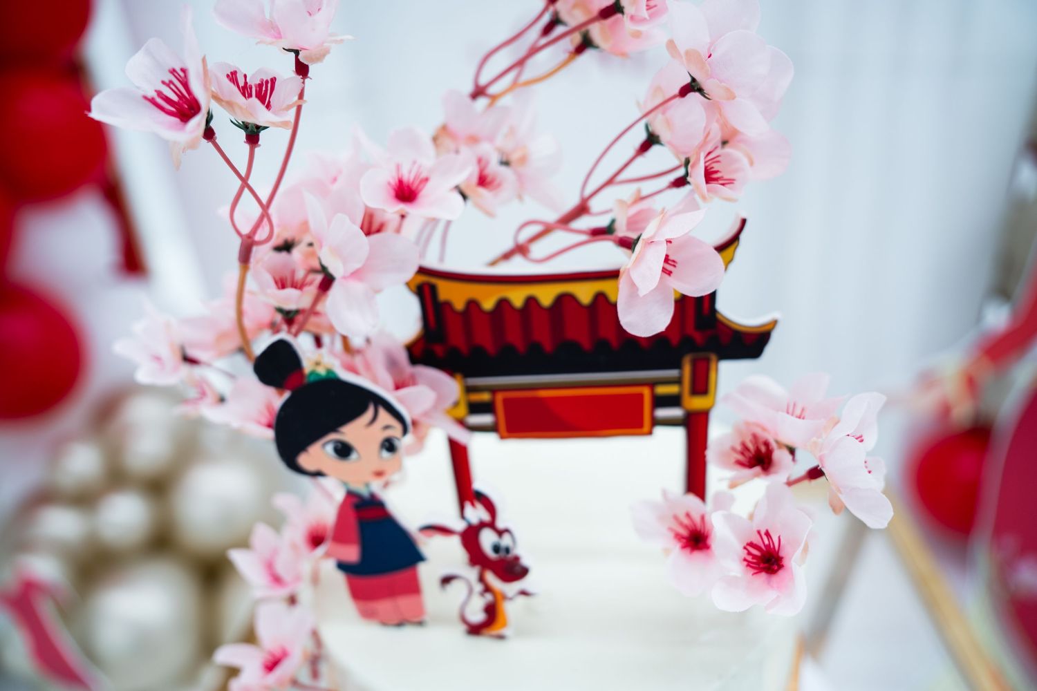 Mulan themed birthday cake with cherry blossom decorations, pagoda topper, and character figurines in red and white.