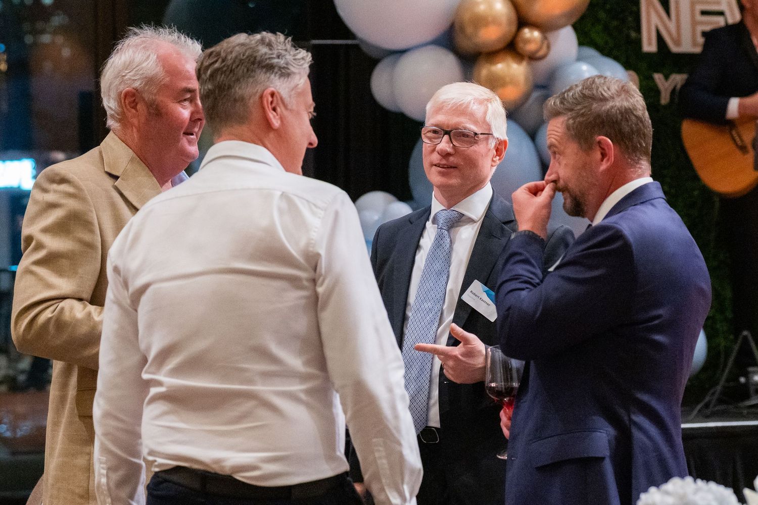 Four men in business attire engaged in conversation at a formal networking event with balloon decorations.