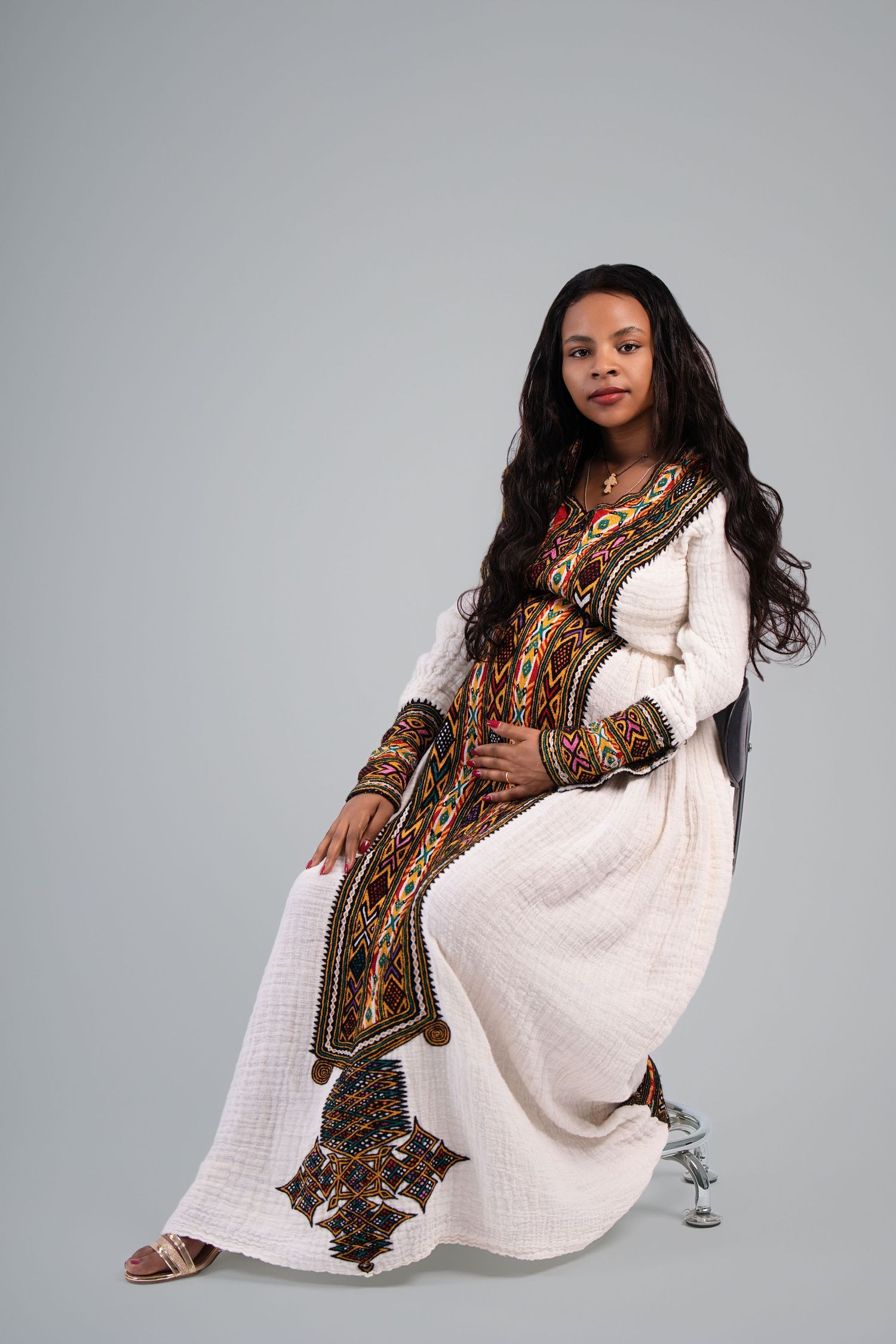 A pregnant woman in traditional Ethiopian white habesha kemis dress with colorful embroidery sits gracefully on a chair.