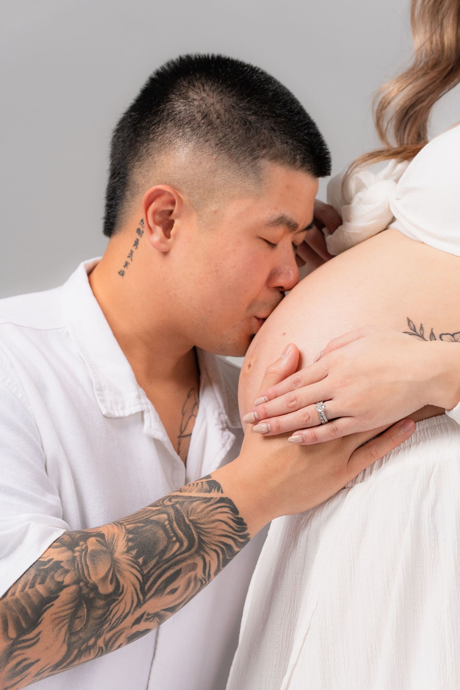 Tattooed man kissing pregnant woman's belly, couple sharing tender moment in white outfits against grey background.