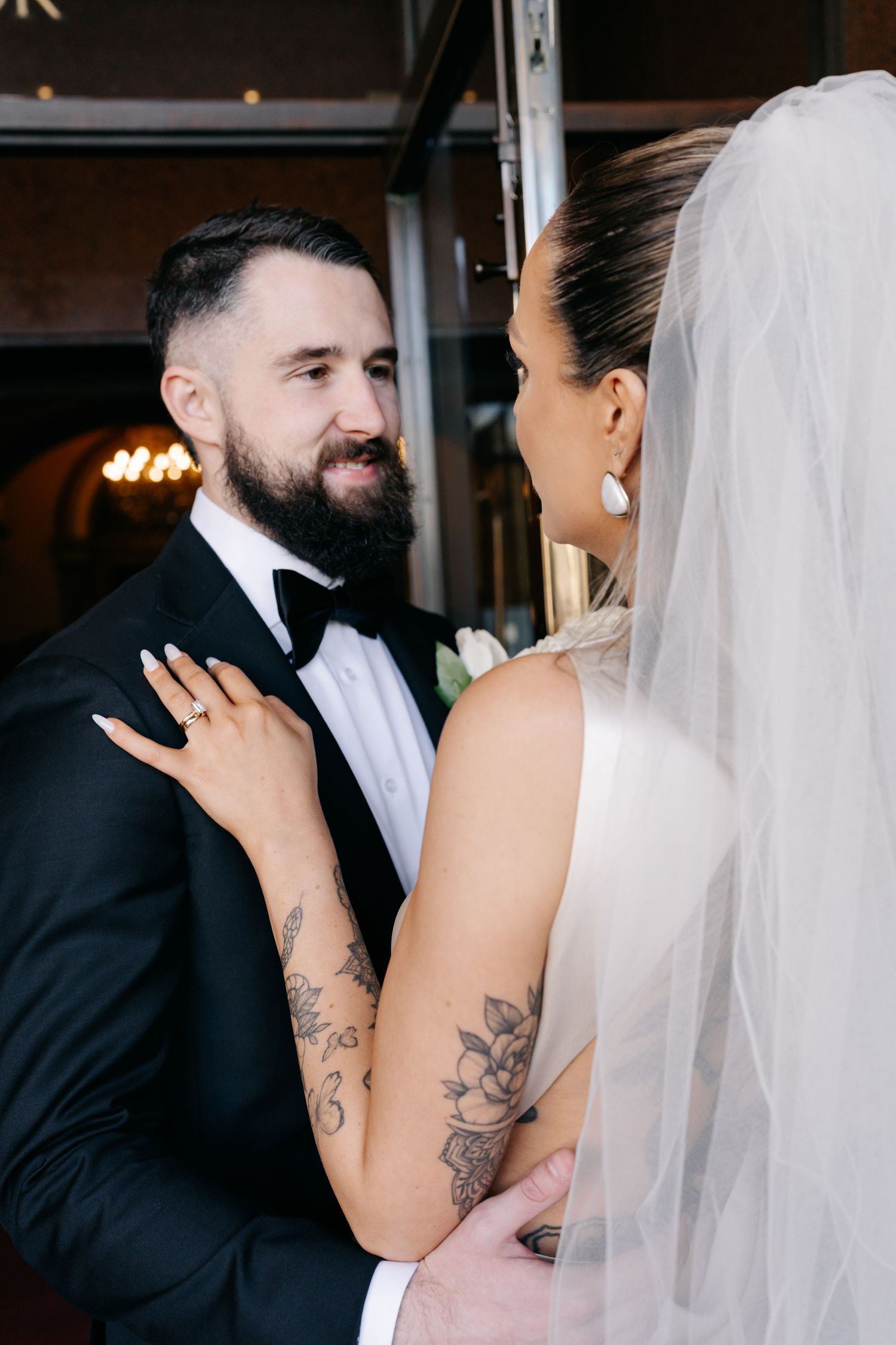 A tender moment between newlyweds showing the bride's floral arm tattoo and groom in classic black tuxedo.