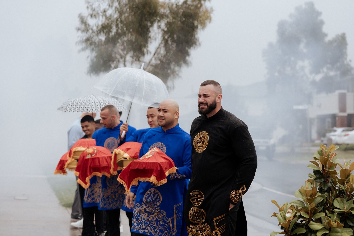 Men in blue Vietnamese ao dai carry red trays in foggy outdoor wedding procession.