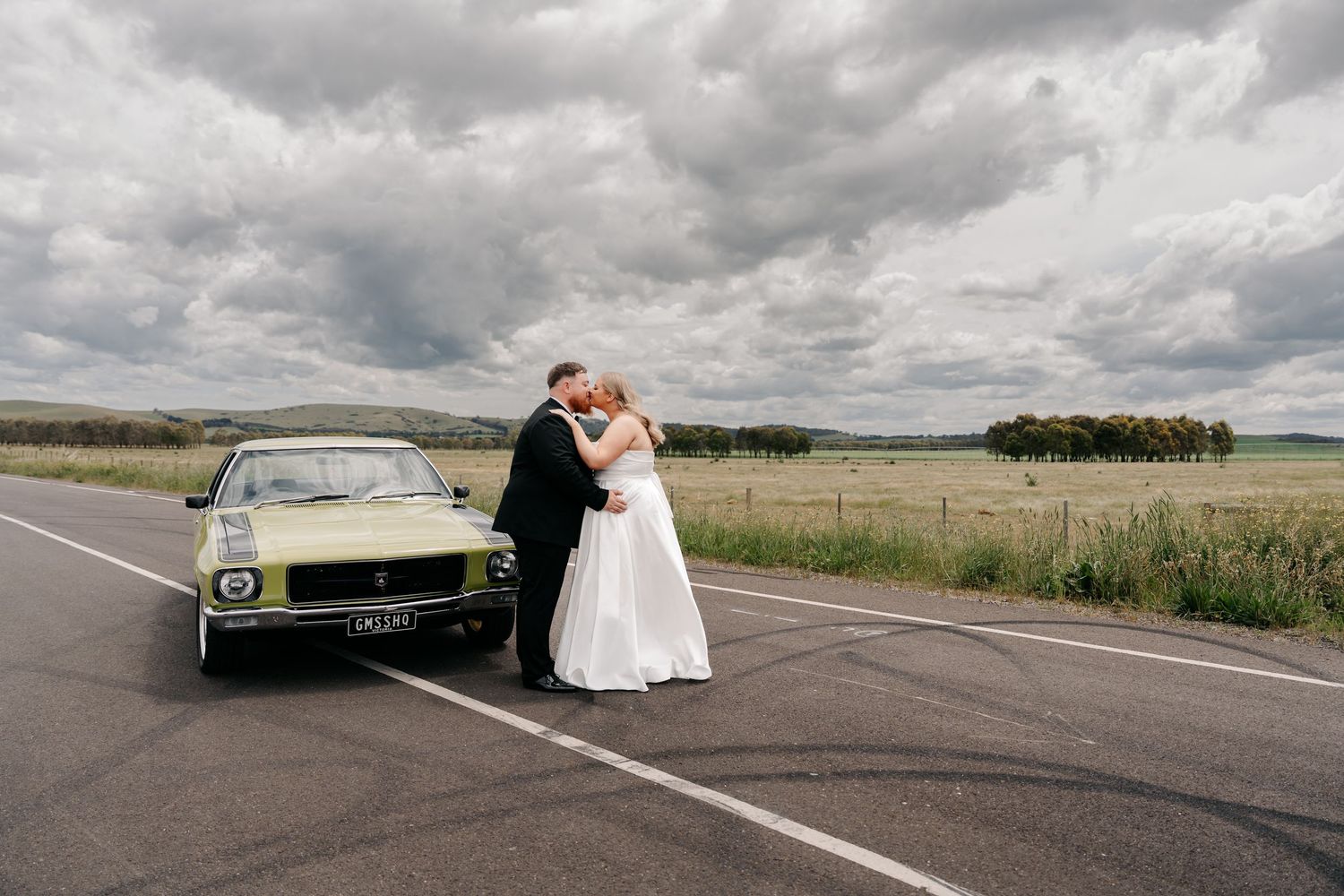 Bride and groom kissing beside a vintage yellow car on a rural road under dramatic cloudy skies.