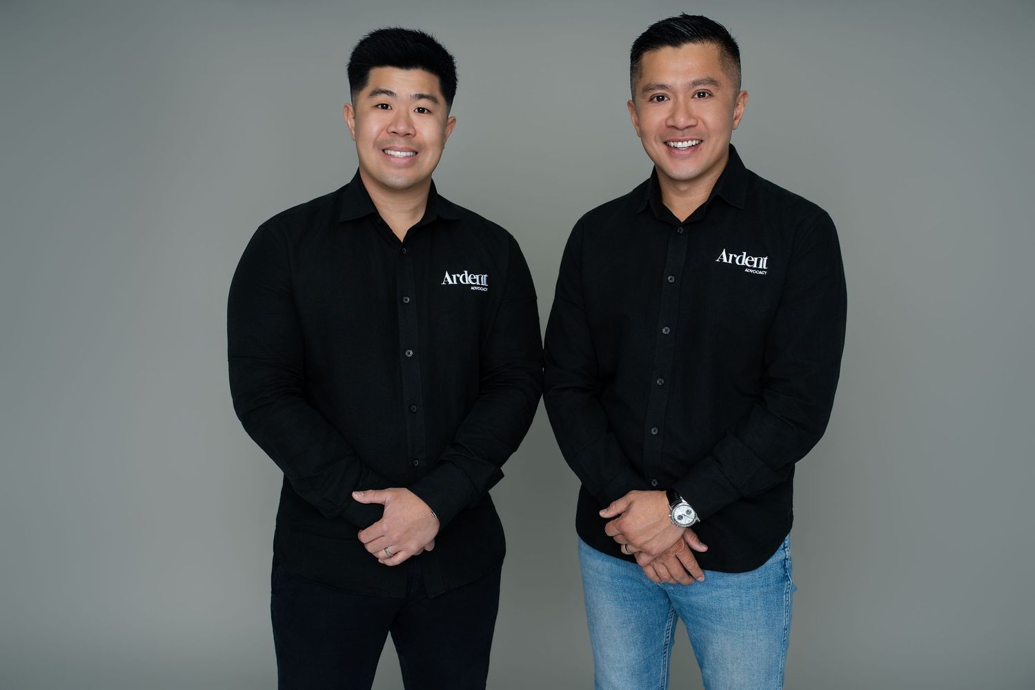 Two men in matching black Ardor branded shirts smiling, posing together against a gray background.