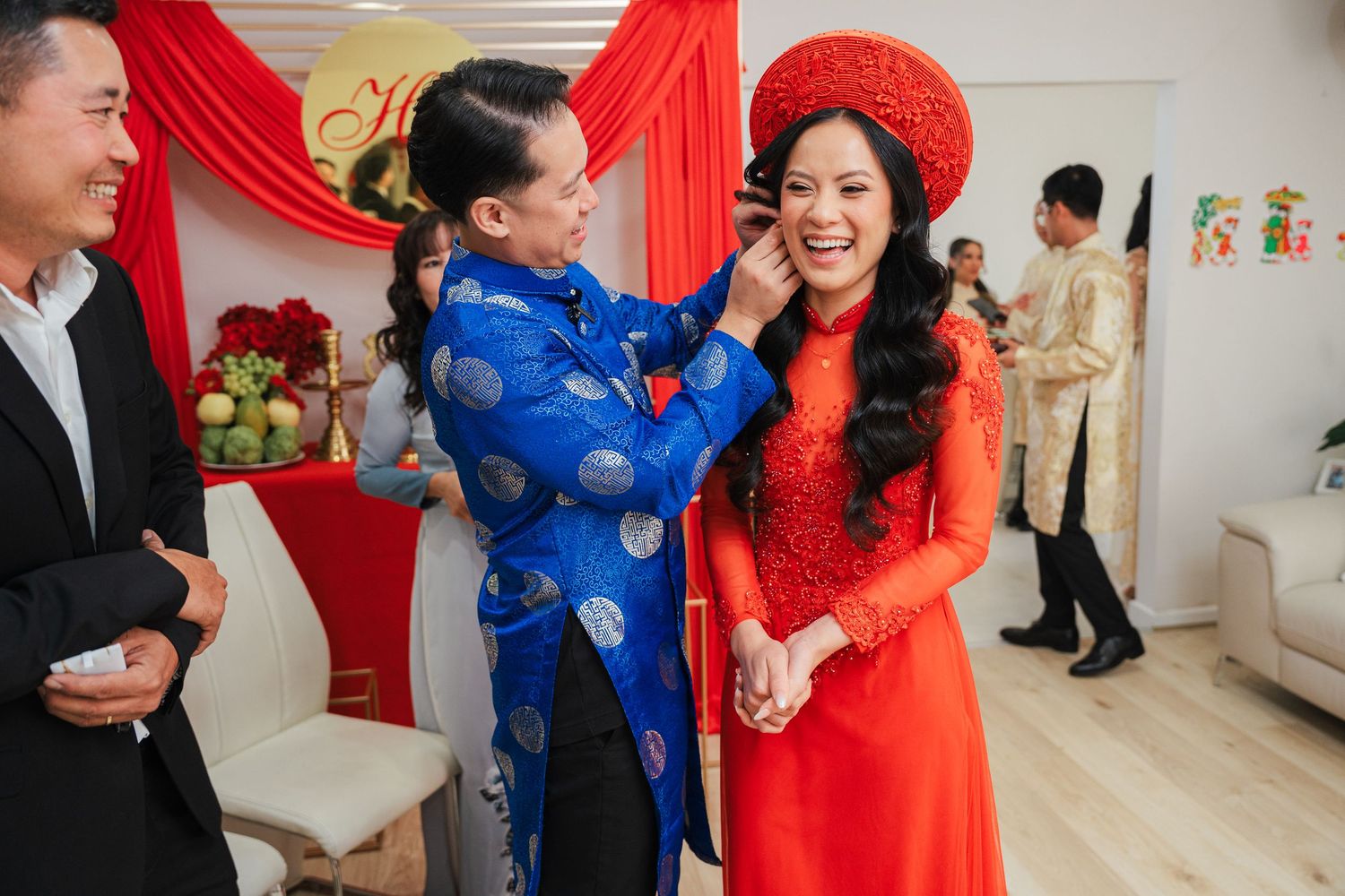 Joyful Vietnamese bride in red ao dai and headdress receiving jewelry from family member at traditional wedding ceremony.