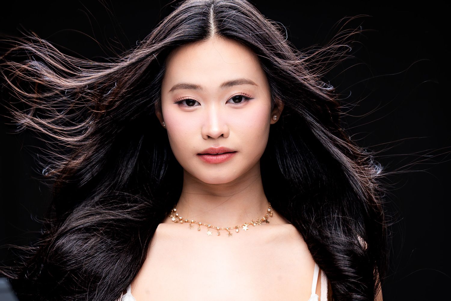 Close-up of young Asian woman with flowing black hair and gold necklace against a dark studio background.