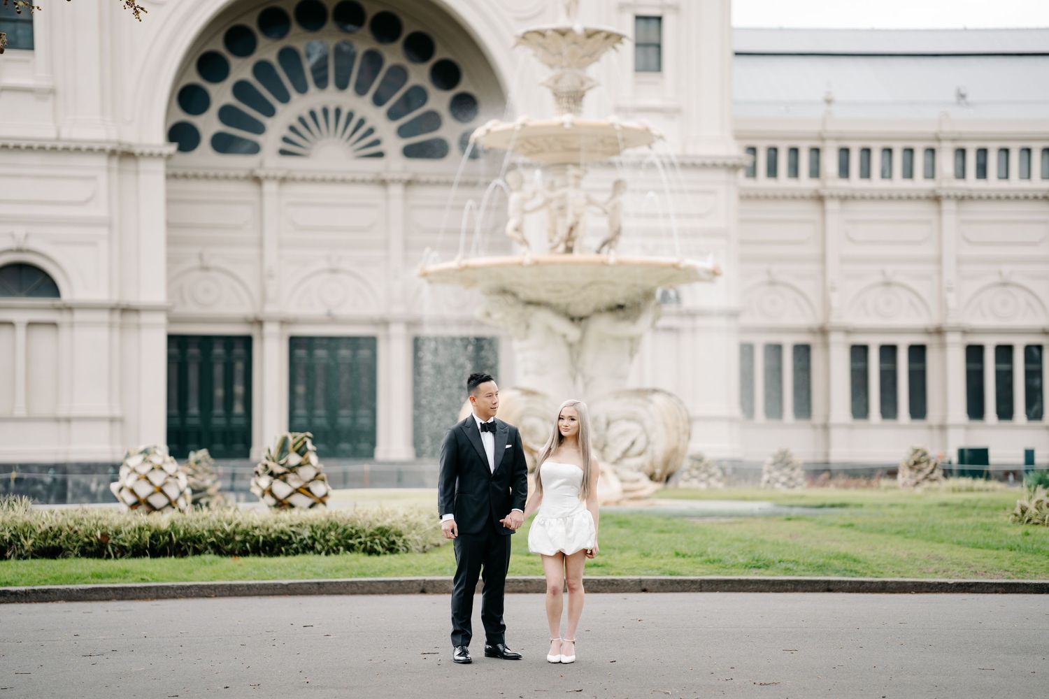 Elegant couple poses in front of ornate architectural building with arched windows.