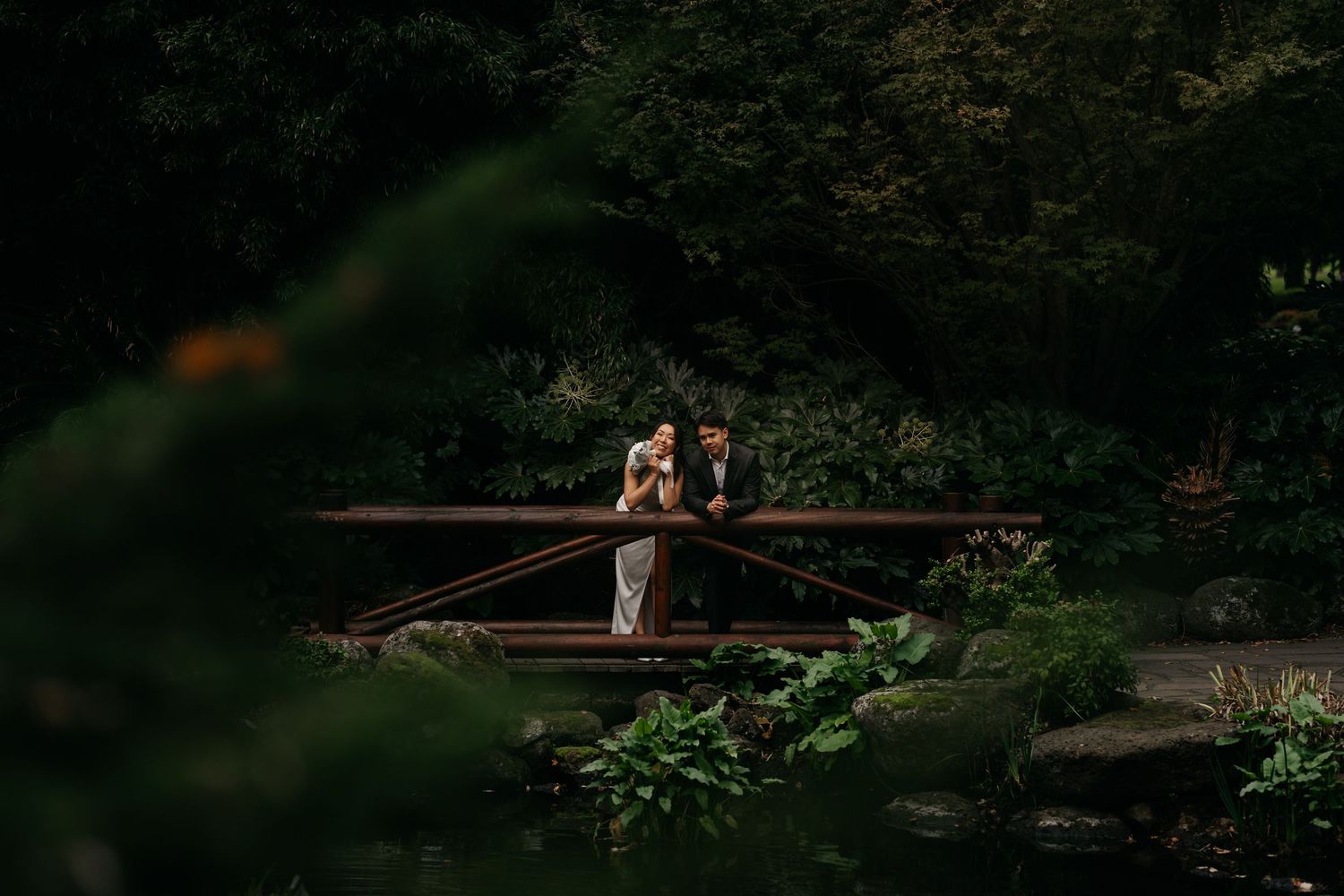 Couple standing on a wooden bridge surrounded by lush green trees, reflecting in a tranquil pond below.