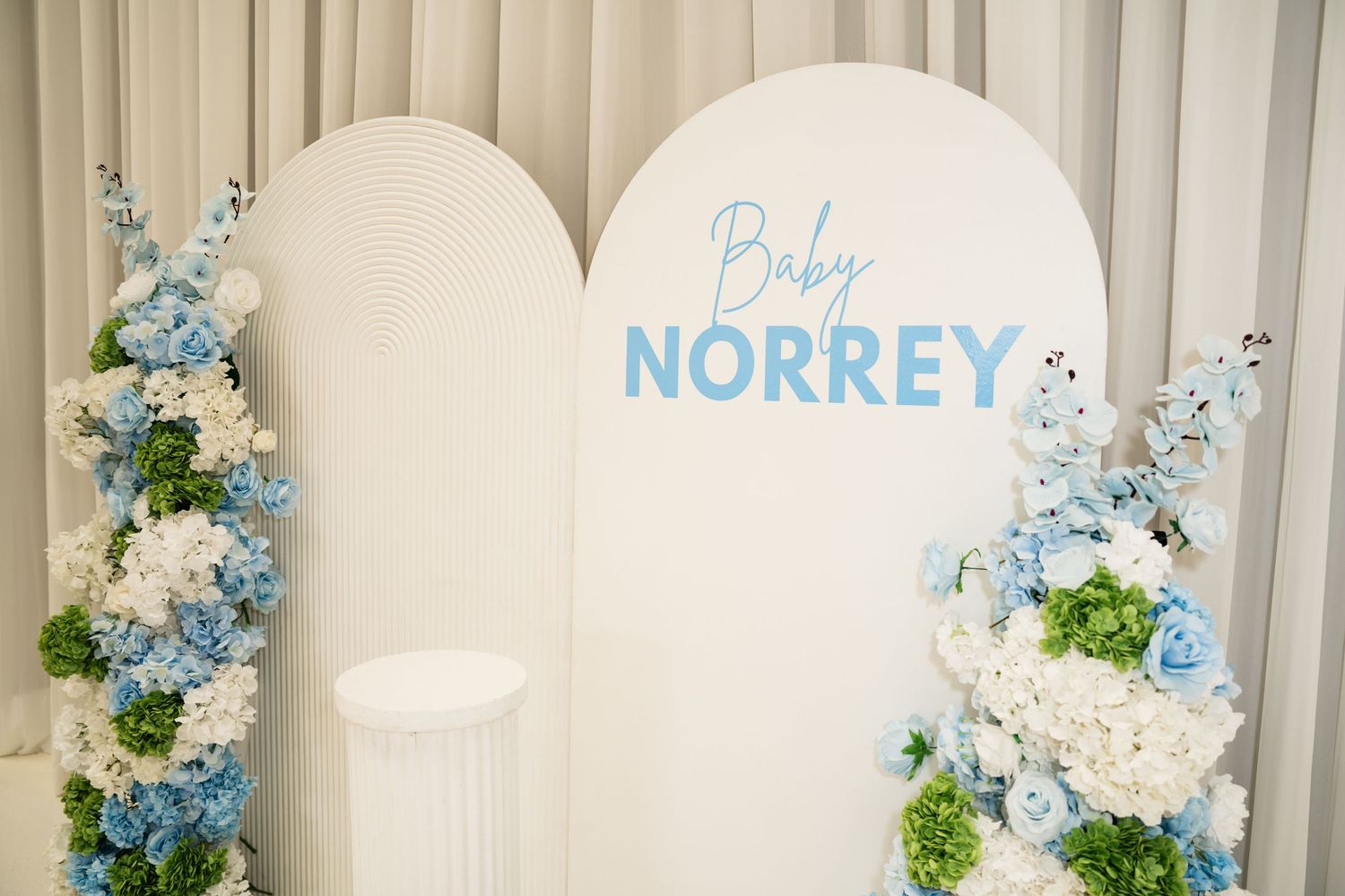 White arch backdrop with blue and white floral arrangements for Baby Norrey baby shower decoration.
