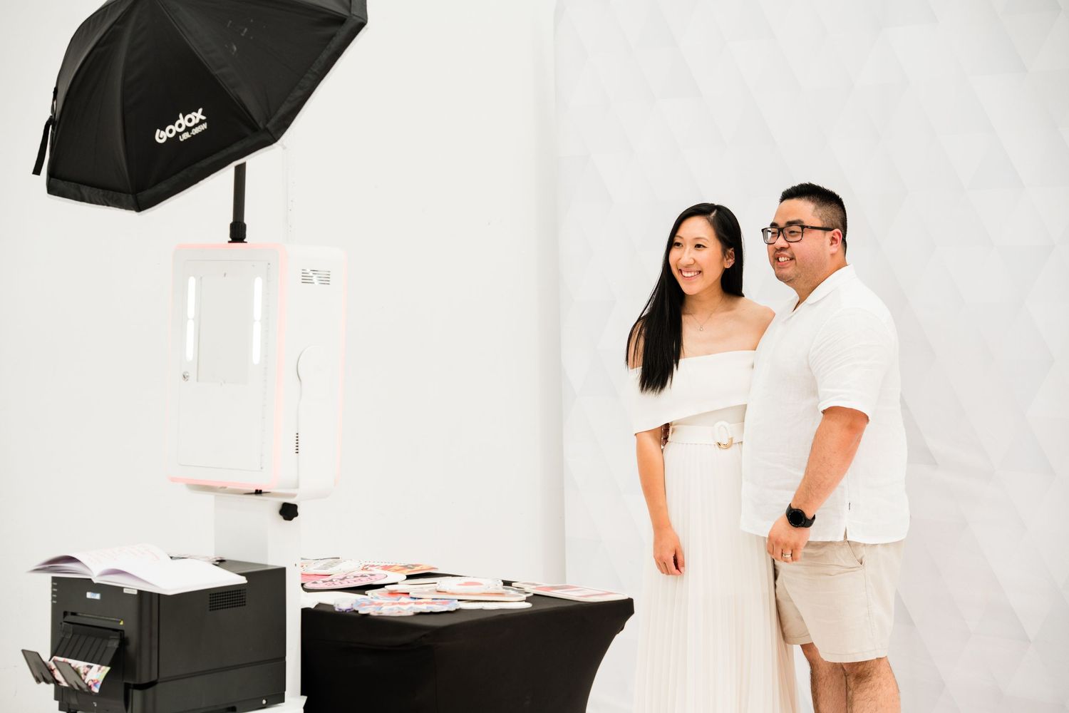 Couple in white outfits posing near a photo booth setup with umbrella light and printer on black table.