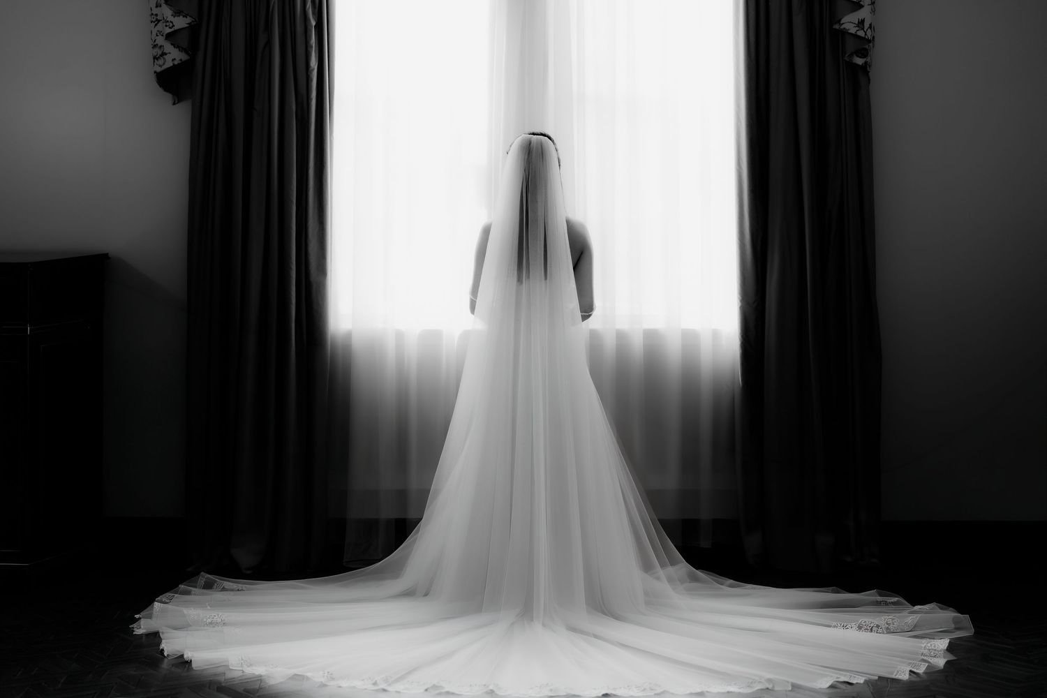 Elegant bride in long veil and white gown stands before sunlit window in black and white photo.