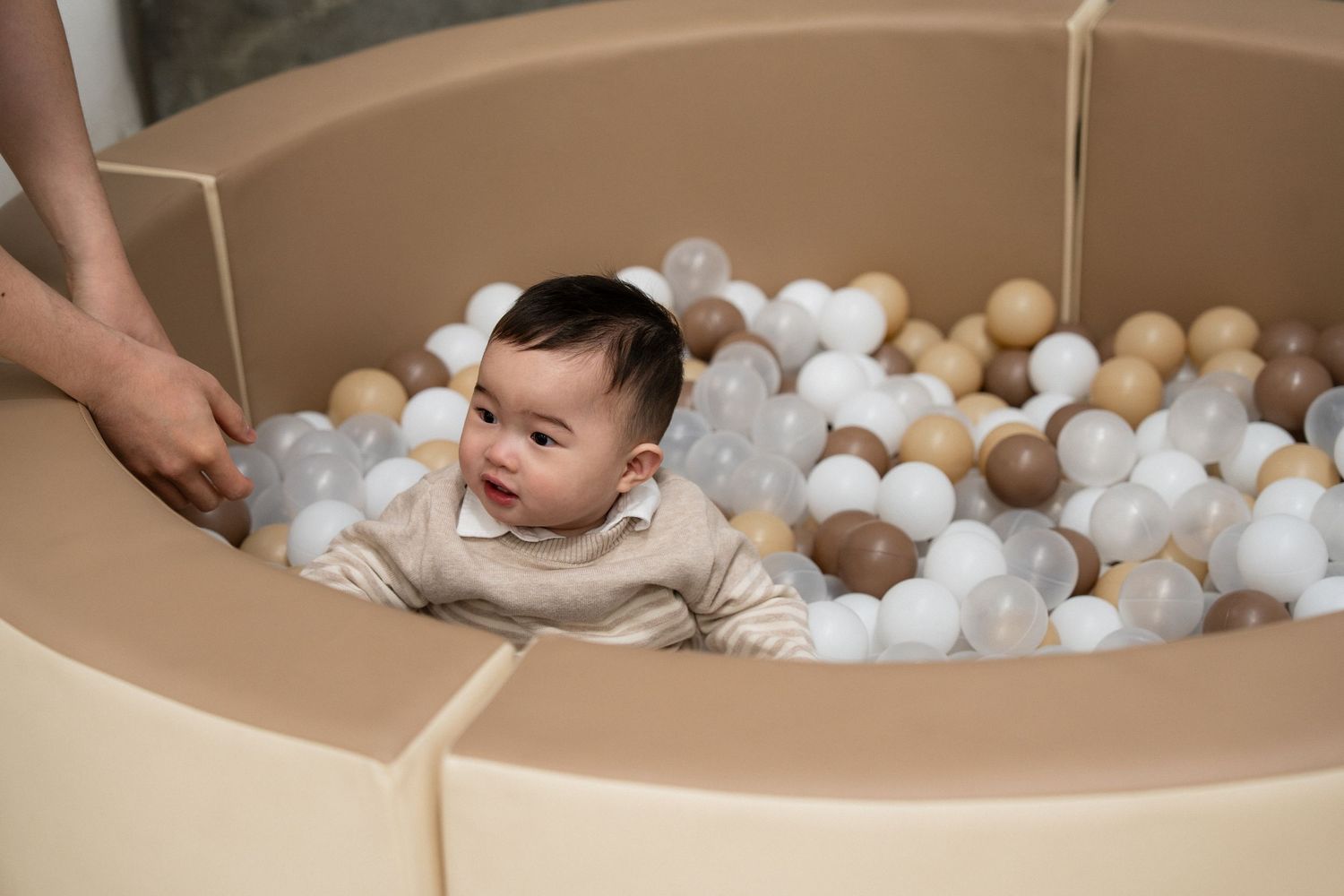 Smiling baby sitting in a beige foam ball pit filled with white, clear, and tan balls, held by an adult.