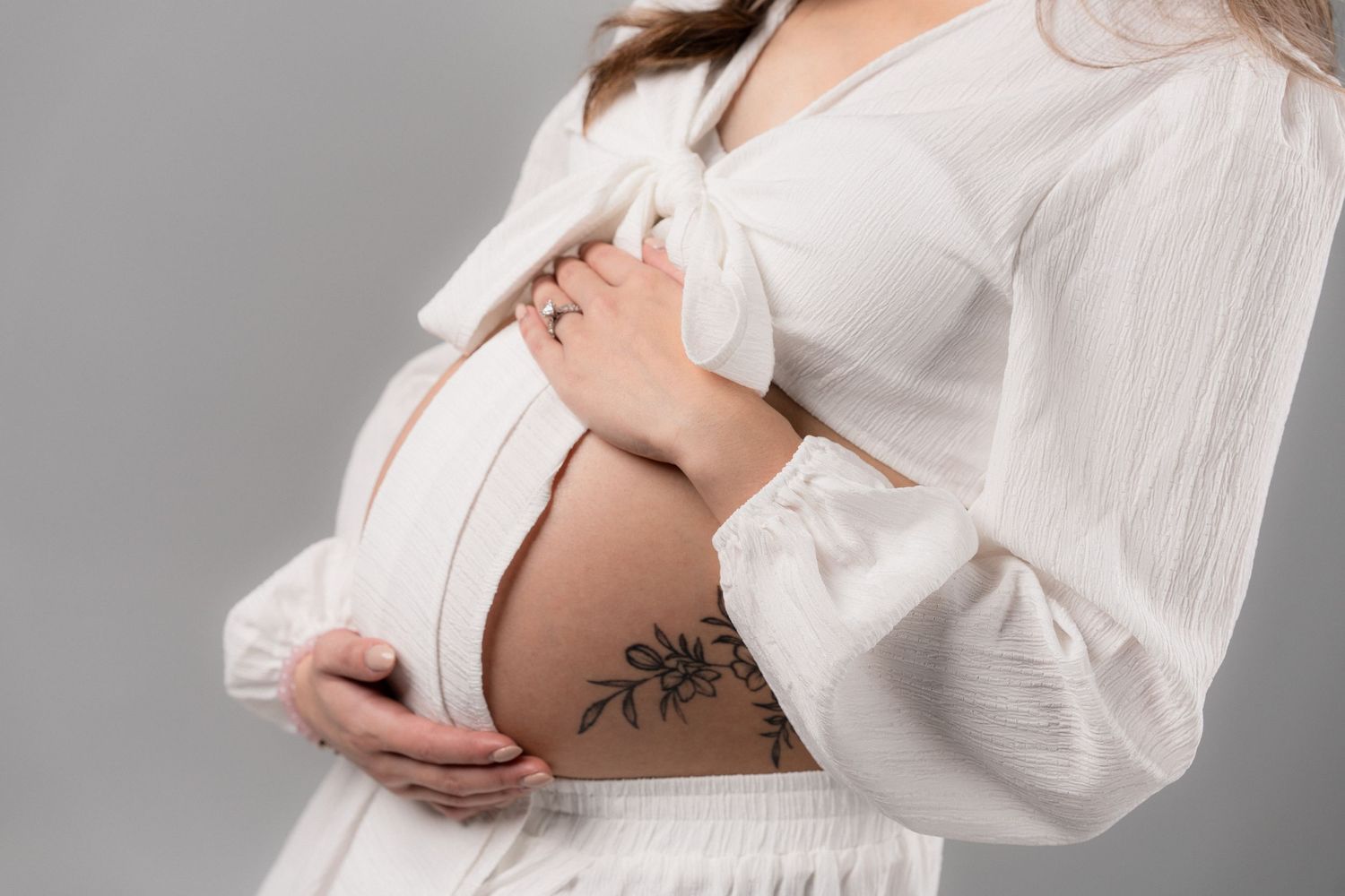 Pregnant woman in white outfit exposing tattooed baby bump, holding belly with both hands on gray background.