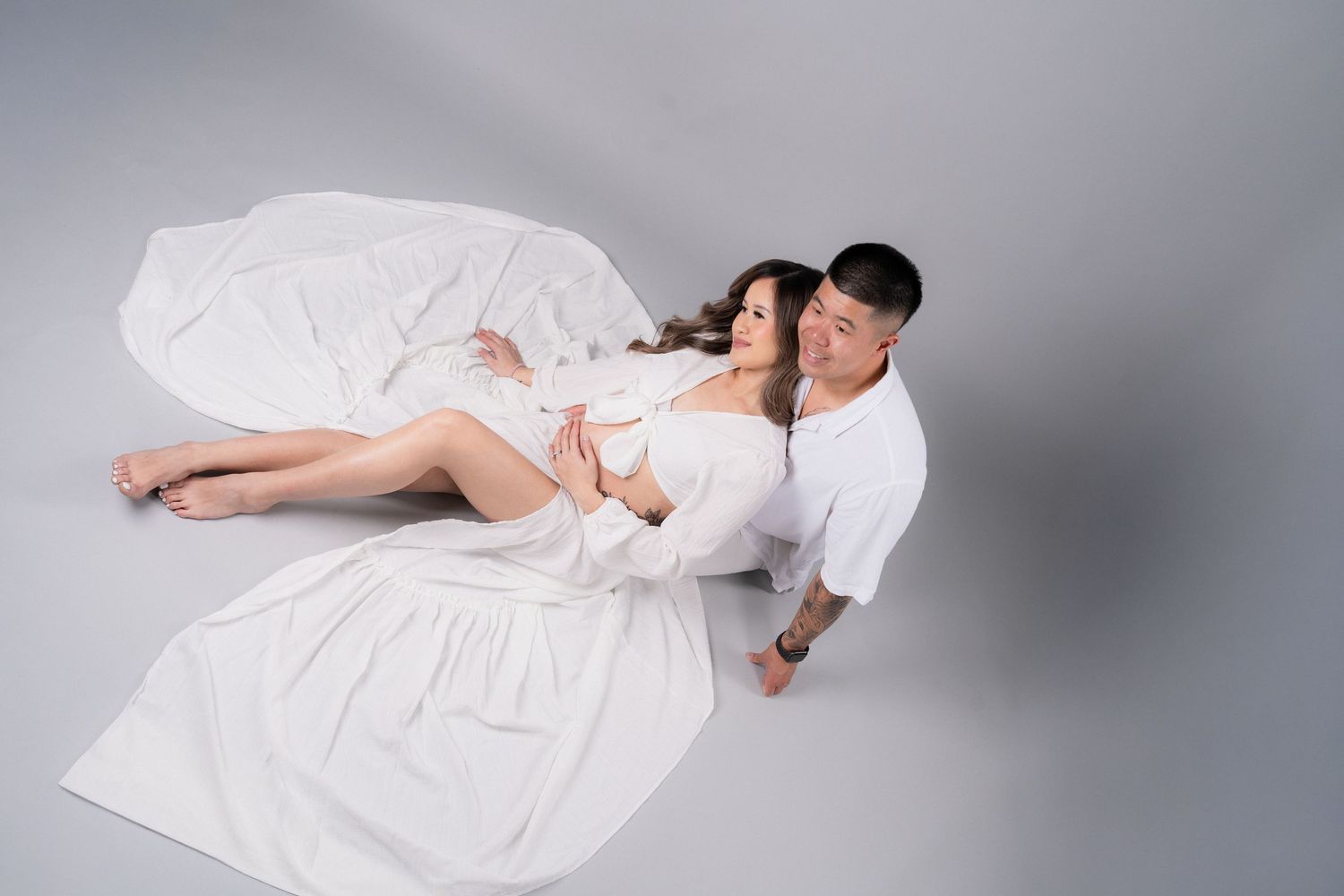 Couple in white outfits posing on floor, pregnant woman cradled by partner in a bright studio setting.