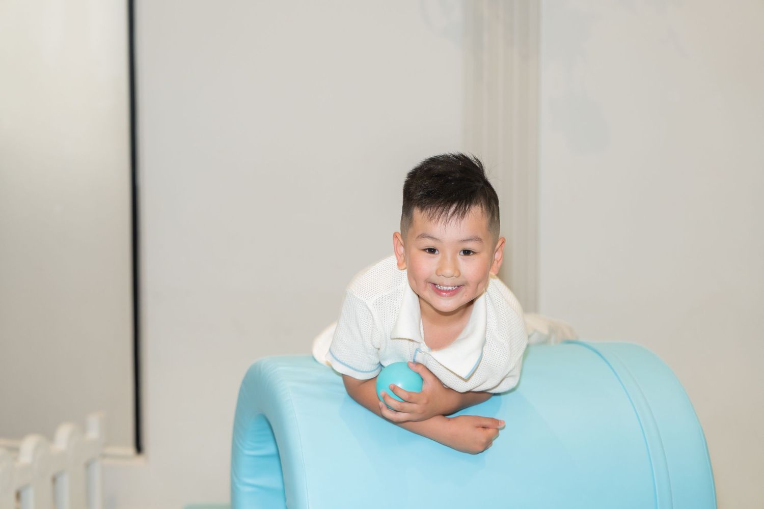 Happy Asian boy playing on a blue foam roller tunnel in a bright indoor play area.