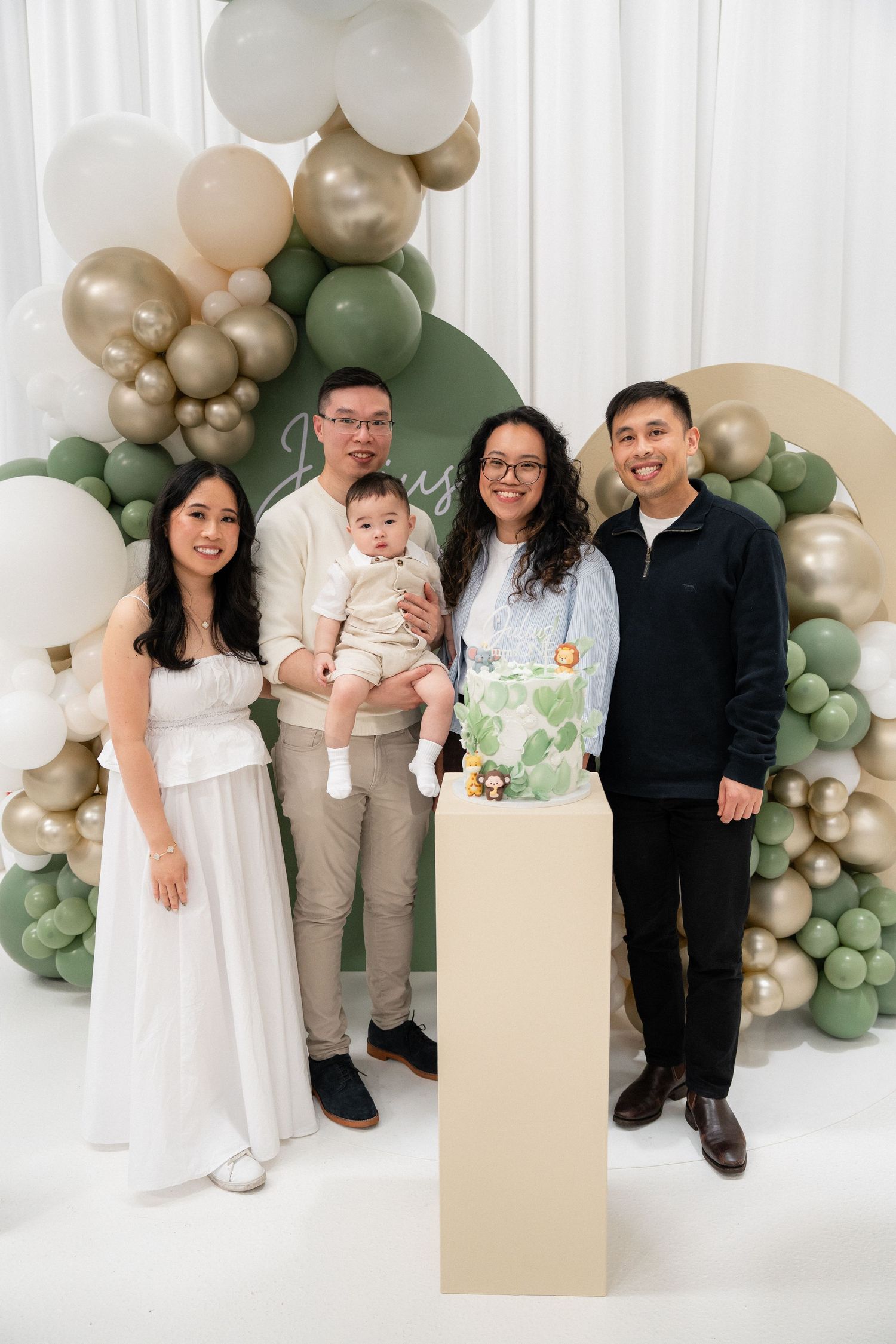 Family posing together at a baby's first birthday party with green and gold balloon arch and decorated cake.
