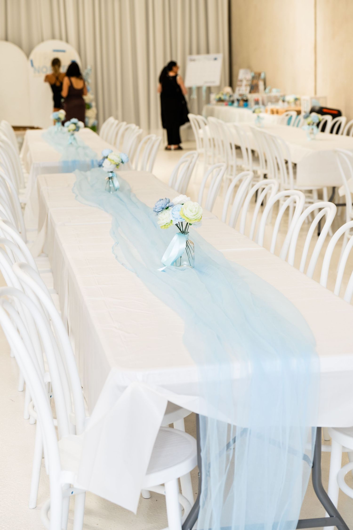 Elegant event setup with long white tables, light blue tulle runner, white chairs, and small floral centerpieces in a bright venue.