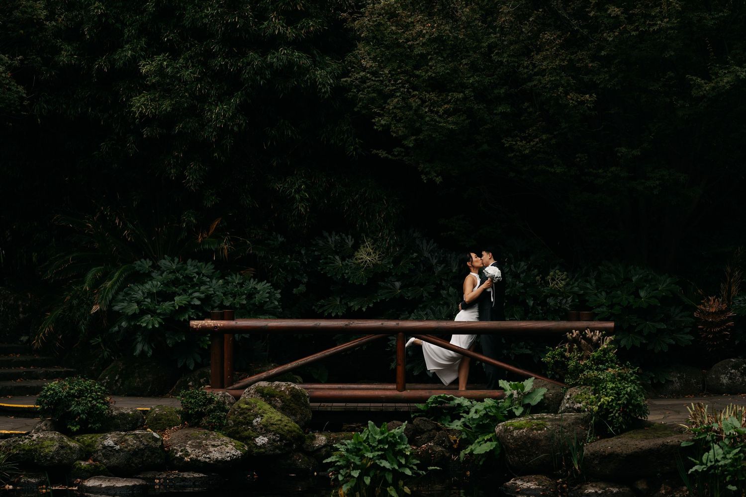 Couple sharing a romantic kiss on a wooden bridge surrounded by lush green forest in a dreamy wedding photo.