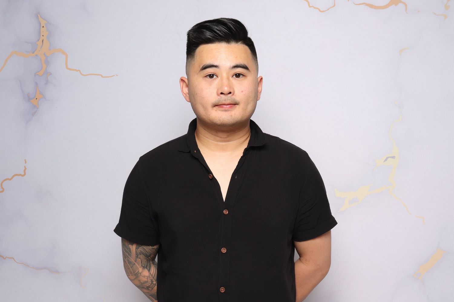 Person wearing a black short-sleeved button-up shirt poses against a marble-textured background.