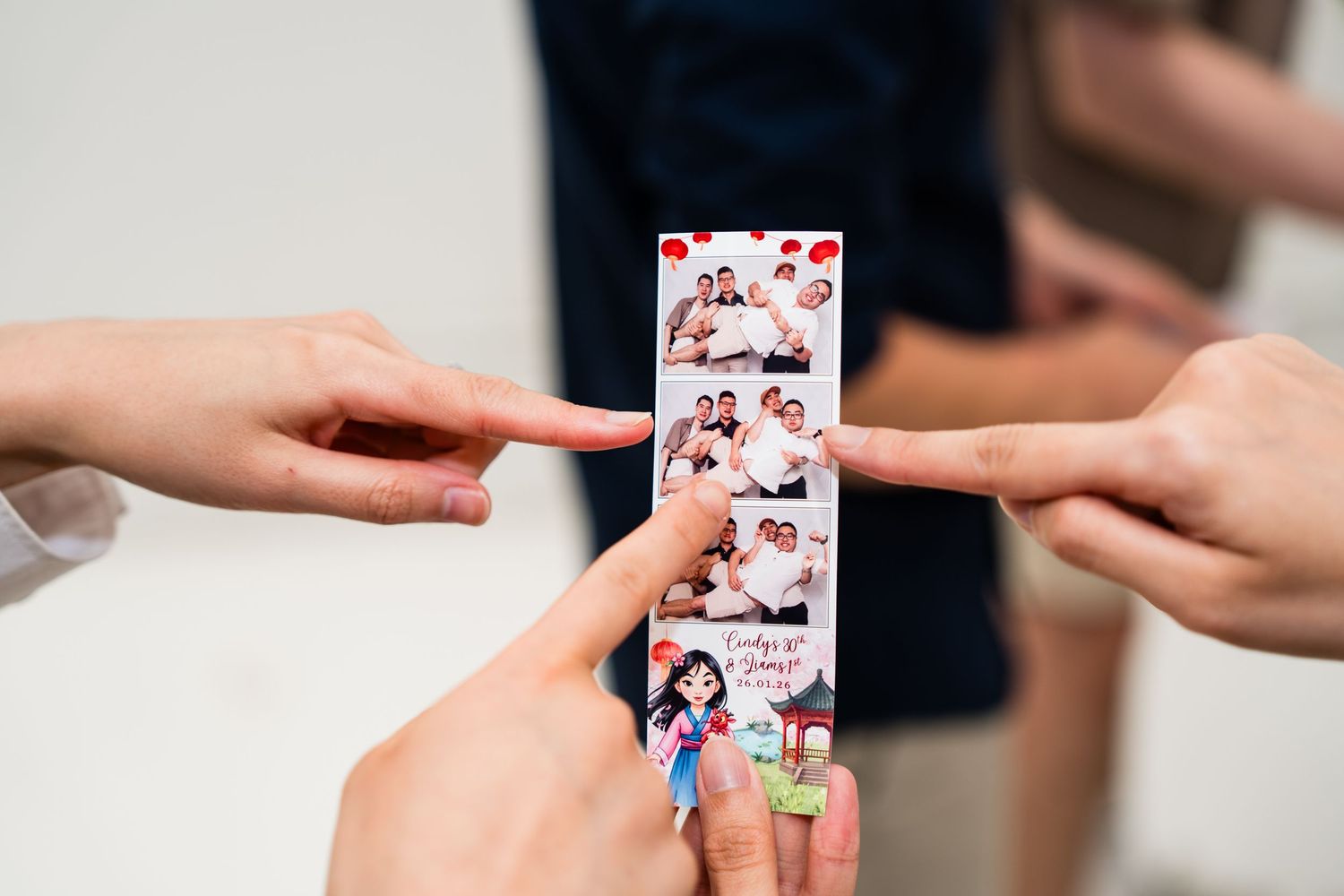 Multiple hands pointing at a photo booth strip showing group photos, held together at a social event.