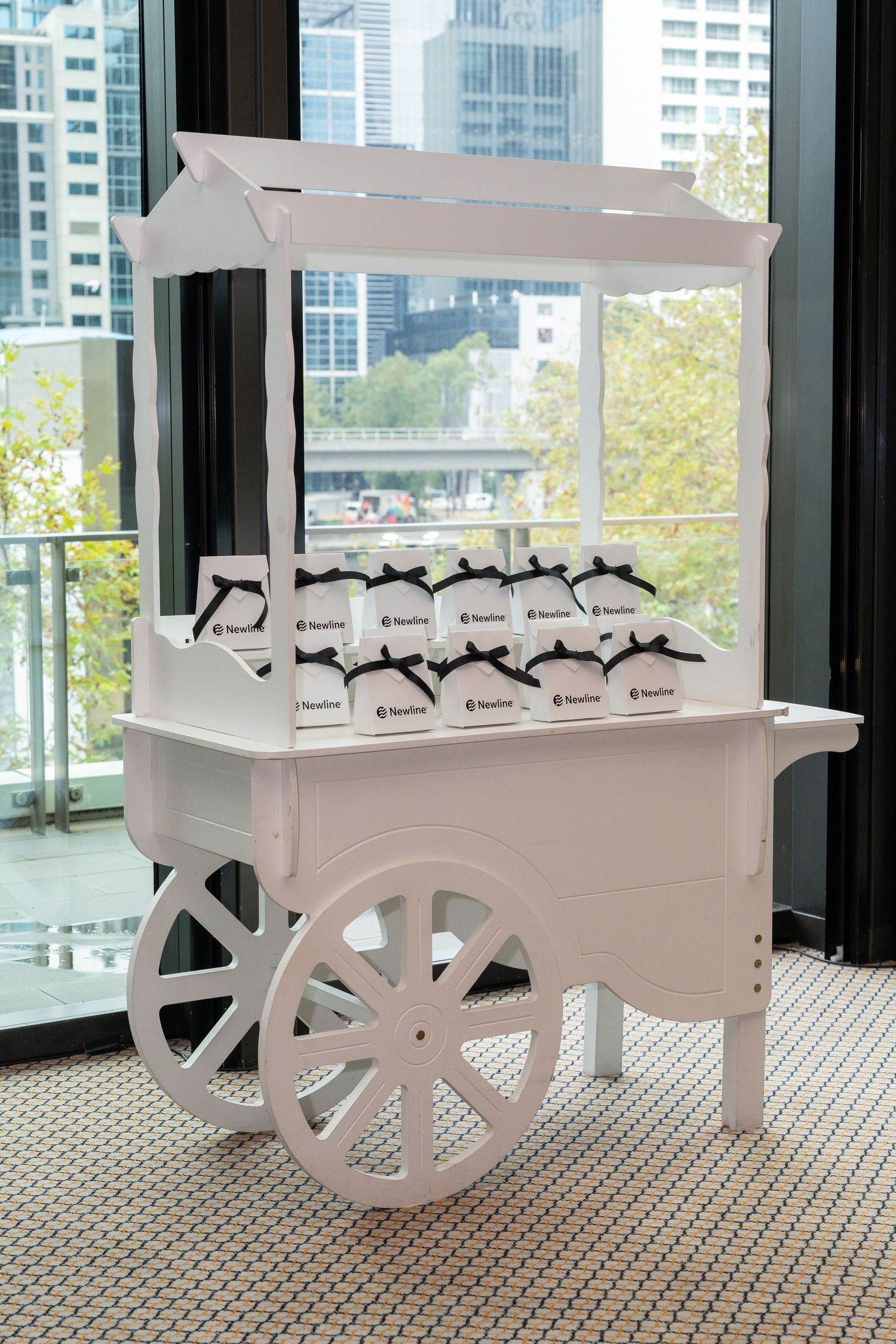 White vintage candy cart displaying branded gift bags with black ribbons, set against a city skyline backdrop.